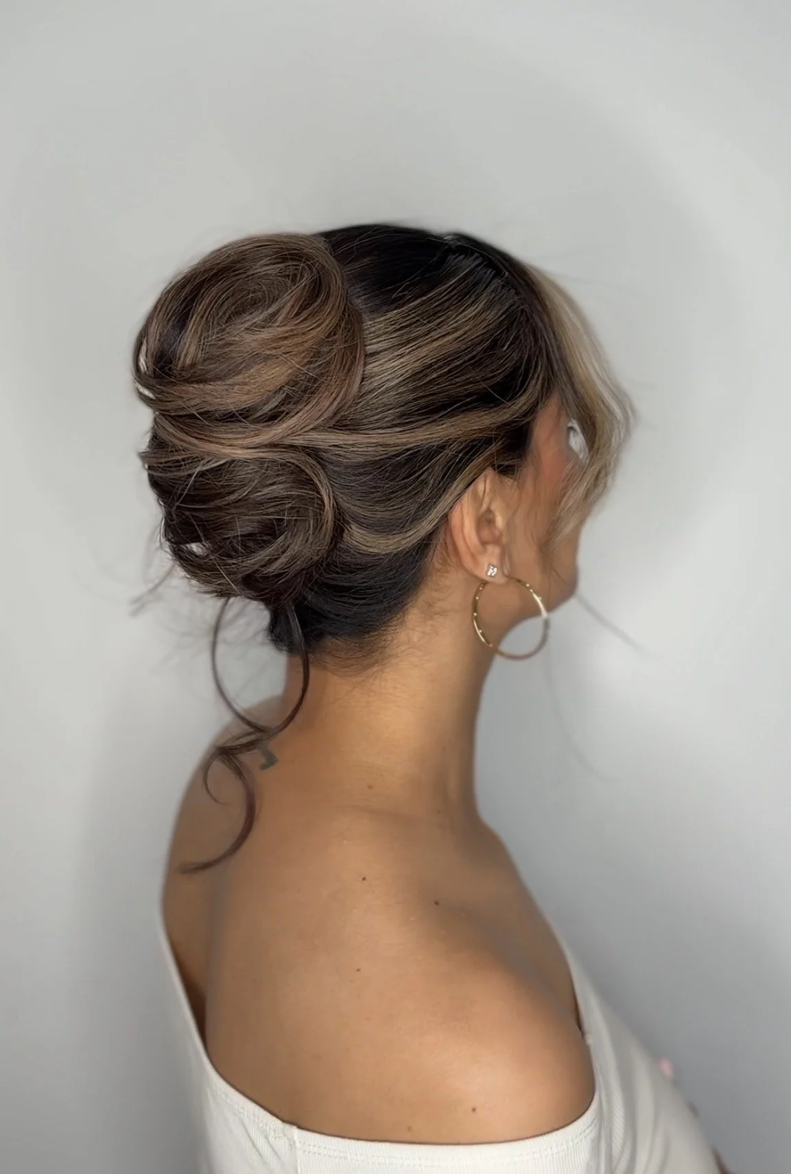 Side view of a woman with an elegant updo hairstyle, featuring styled brown hair with blonde highlights, wearing hoop earrings and a white off-the-shoulder top.