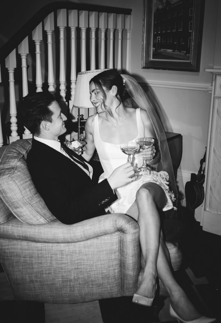 A bride and groom sitting on a sofa, smiling and holding champagne glasses, inside a home with a staircase and framed artwork on the wall.