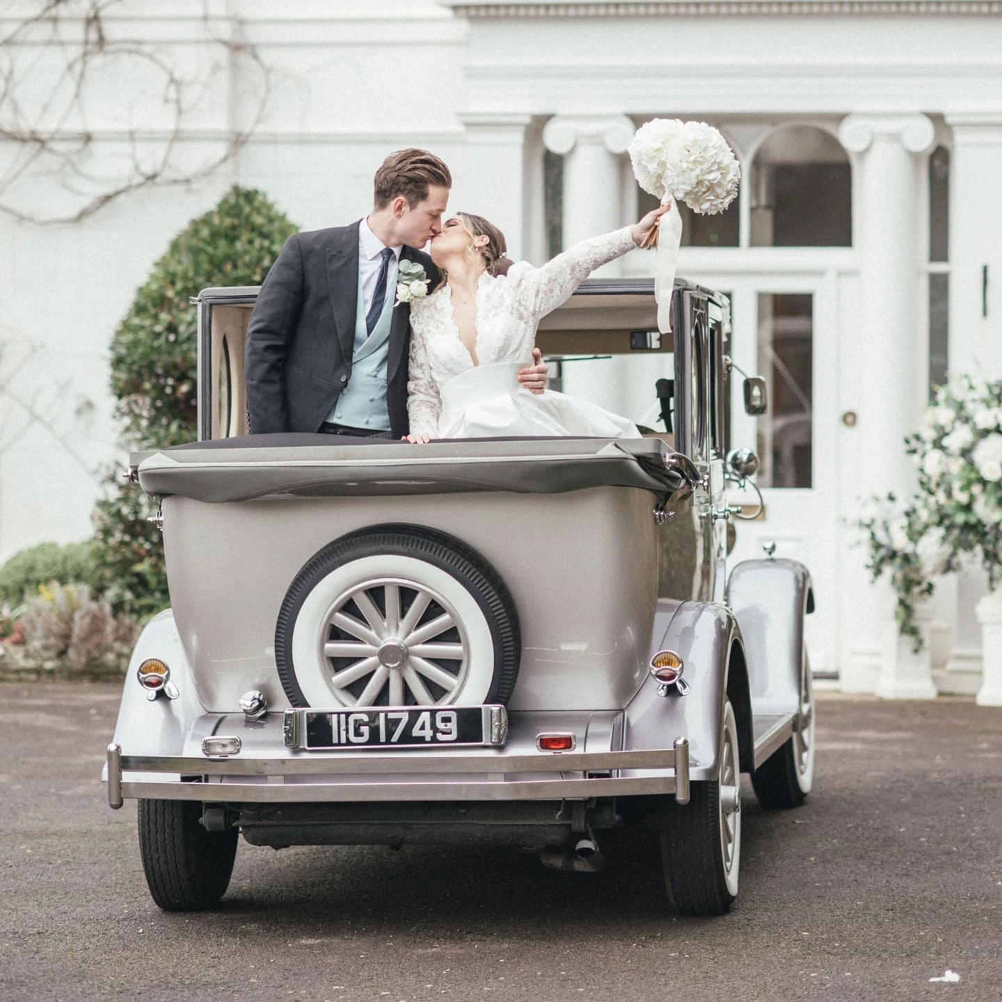 A bride and groom kissing inside a vintage car with a bouquet, in front of a white building with columns and greenery.