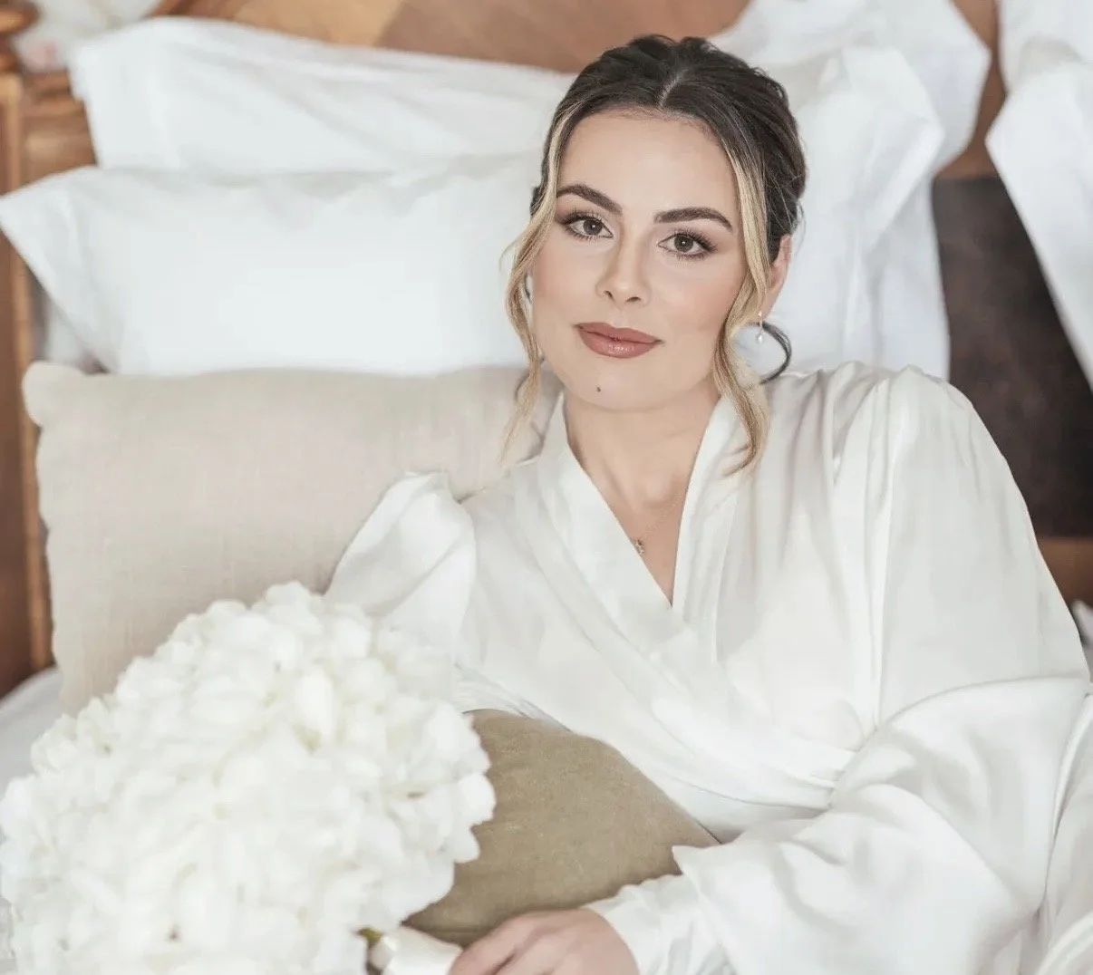 A woman with light skin, brown hair with blonde highlights, wearing a white satin robe, lying on a bed with white pillows and beige cushions, holding a bouquet of white flowers, in a cozy bedroom setting.