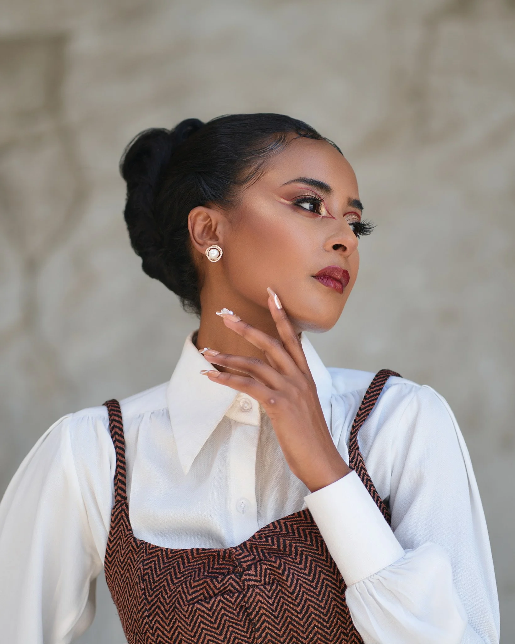 A woman with styled black hair, wearing pearl earrings, bold makeup with winged eyeliner and red lipstick, a white shirt with a Victorian collar, and a brown and black chevron-patterned vest, poses in front of a blurred neutral background.