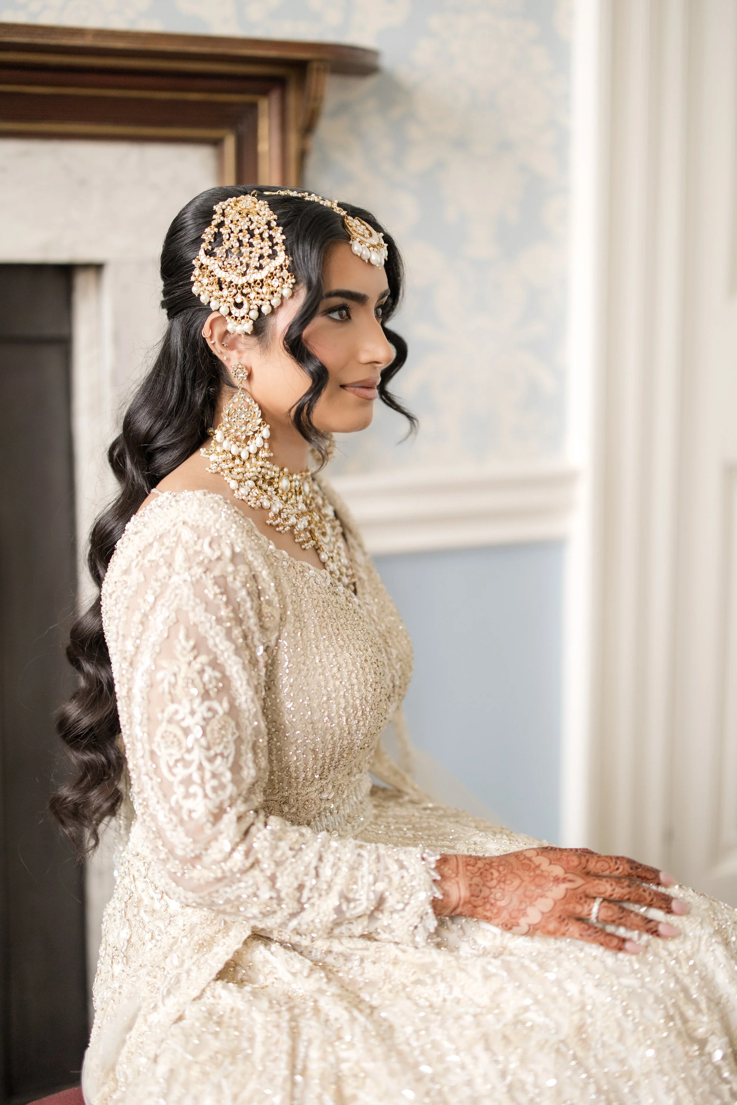A woman dressed in traditional bridal attire, including ornate jewelry, a heavily embroidered beige and gold dress, and intricate henna on her hands, sitting calmly indoors near a fireplace.