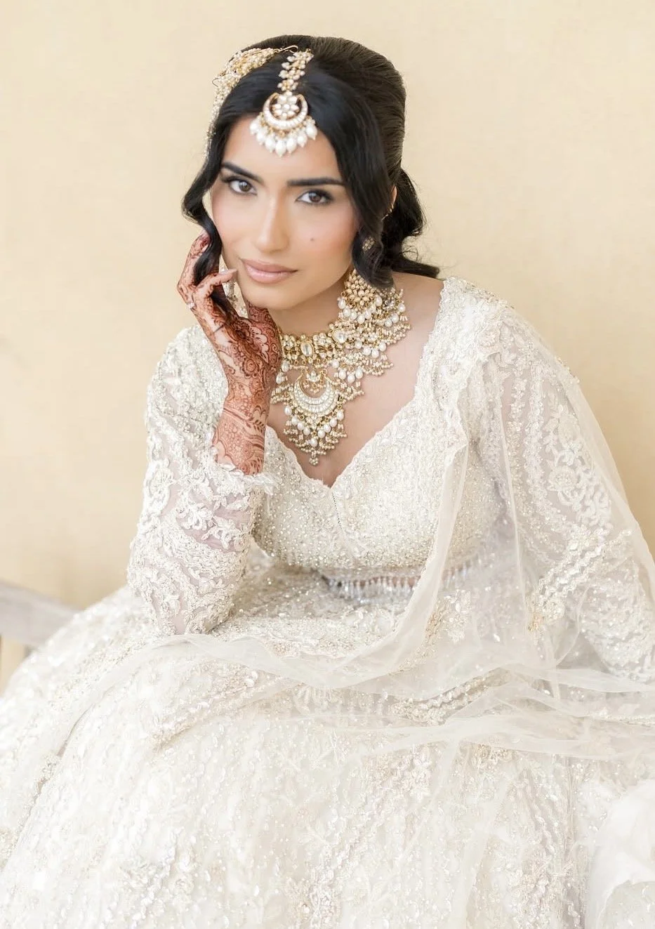 A woman in traditional Indian wedding attire, wearing a cream-colored embroidered dress and heavy jewelry, including a necklace, earrings, and a headpiece, with henna on her hand.