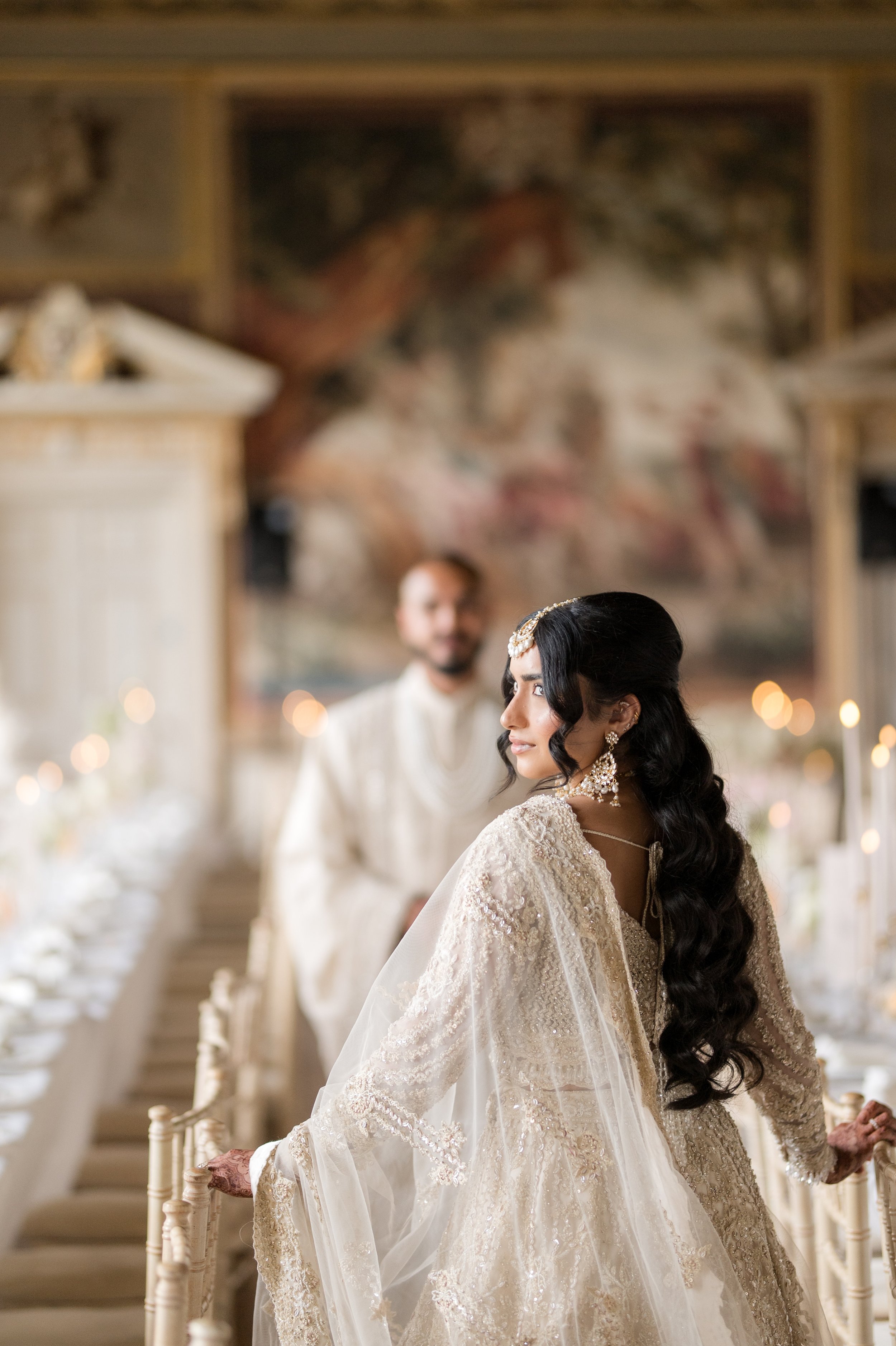 A bride in an ornate cream-colored wedding dress with lace and beading, standing on a staircase during her wedding ceremony, with a groom and guests in the background, inside an elegant venue with wood ceiling beams and subdued lighting.