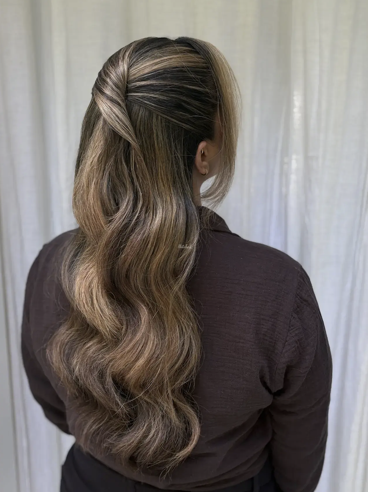 Back view of a woman with long, wavy, light brown hair, styled with loose curls and subtle highlights, wearing a dark brown top.