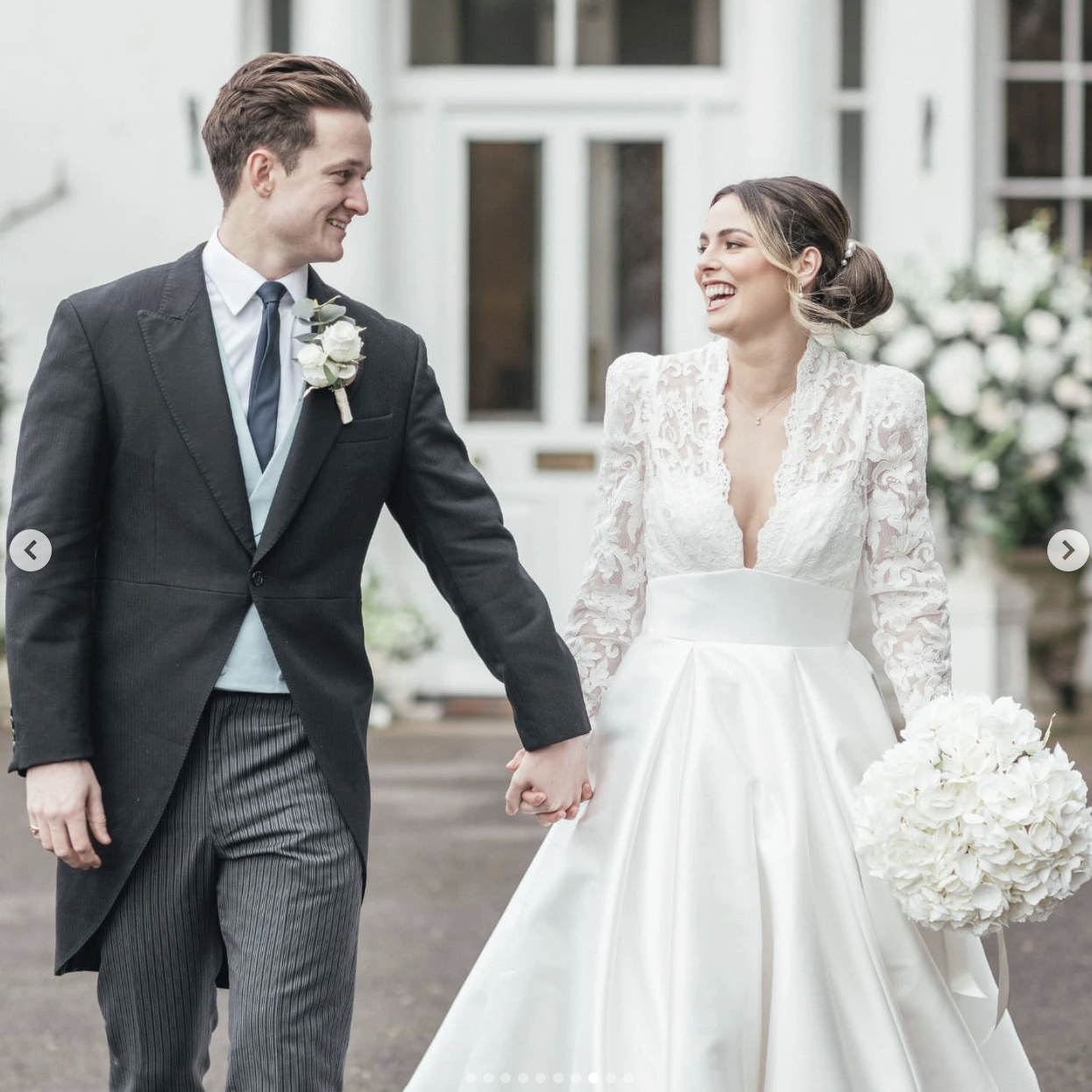 A bride and groom holding hands and smiling at each other outside on their wedding day, with a white building and flowers in the background.