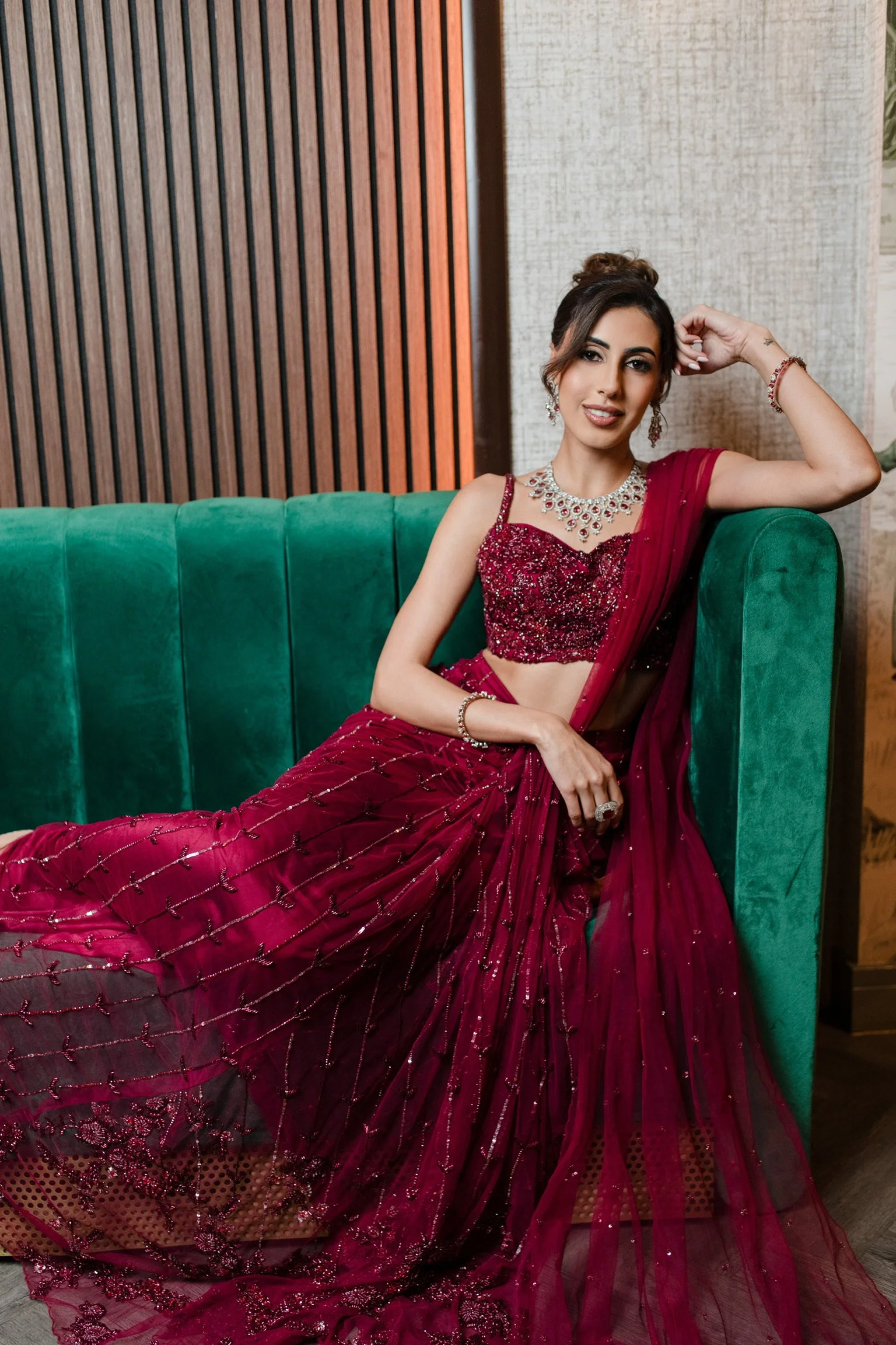 A woman in a deep red, heavily embellished traditional Indian saree, sitting on a green velvet couch with one arm resting on her head, smiling at the camera. She is wearing matching jewelry including earrings, a necklace, bracelets, and a ring, with 
