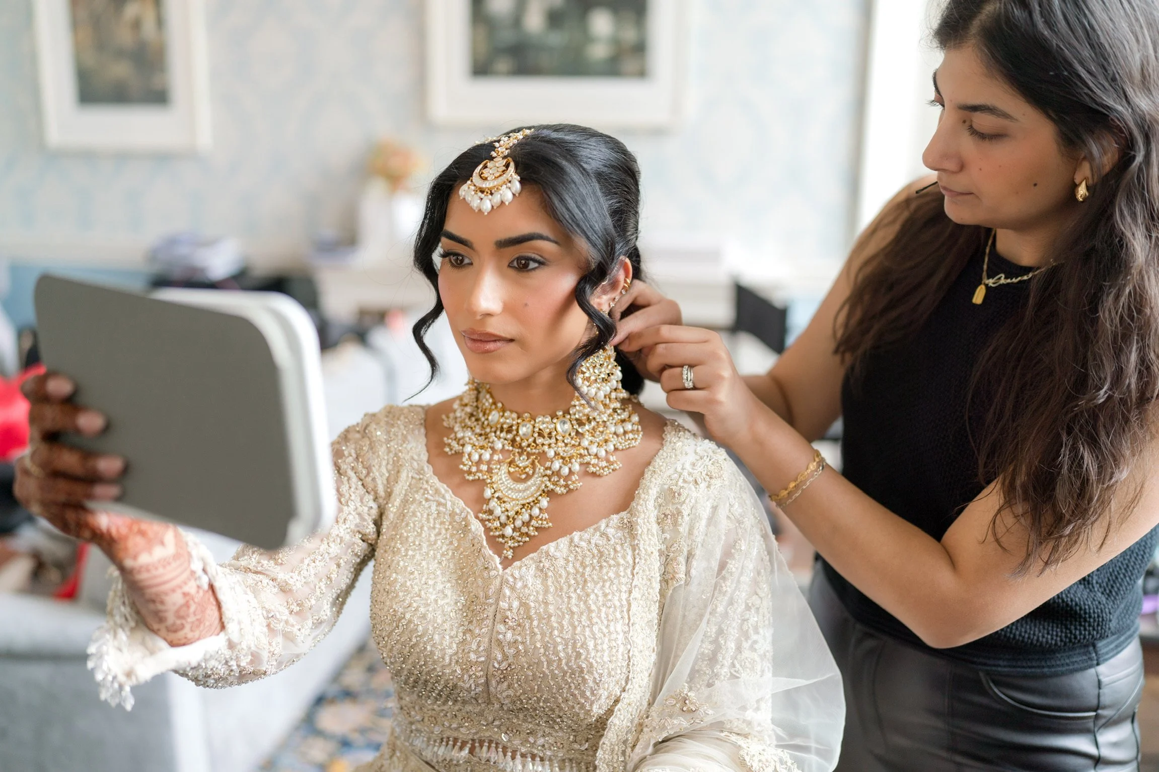 A bride dressed in traditional Indian wedding attire with elaborate jewelry and henna on her hands, taking a selfie while a woman adjusts her earrings in a well-lit room.