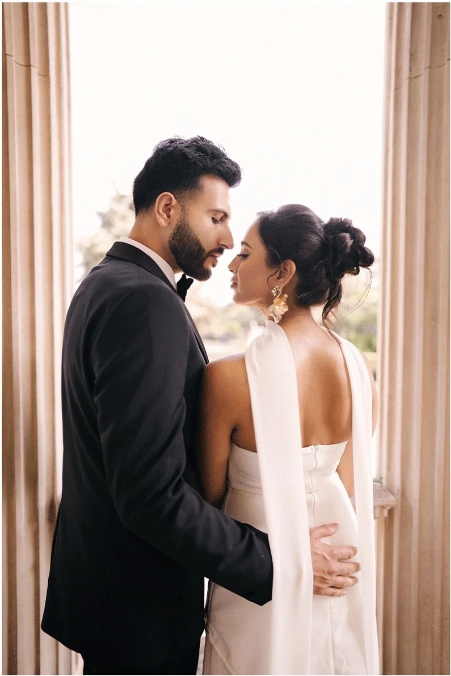 A bride and groom standing close together, looking at each other, with the groom's hand on the bride's waist, inside a room near a window.