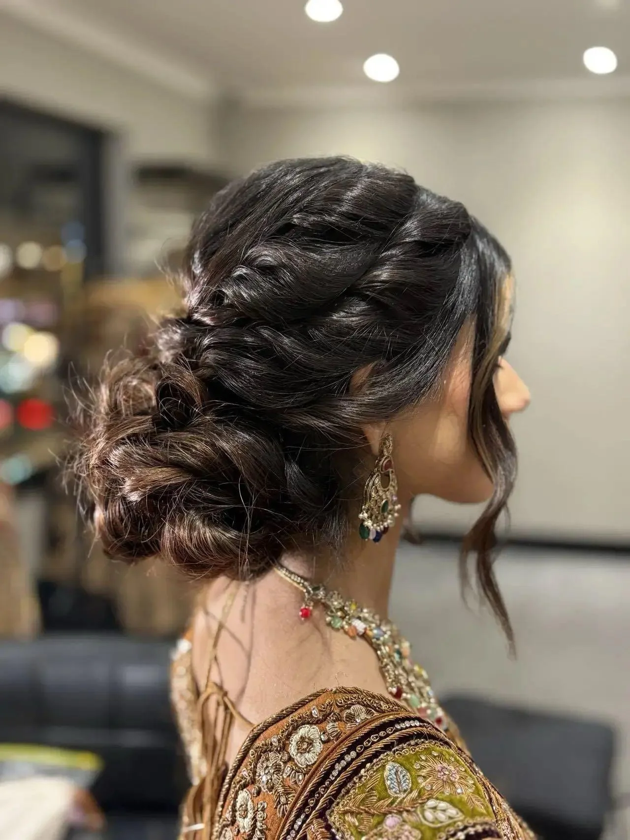 A woman with dark, wavy hair styled in an elegant updo with loose curls framing her face, wearing intricate traditional jewelry and clothing, indoors with a neutral background and bright ceiling lights.