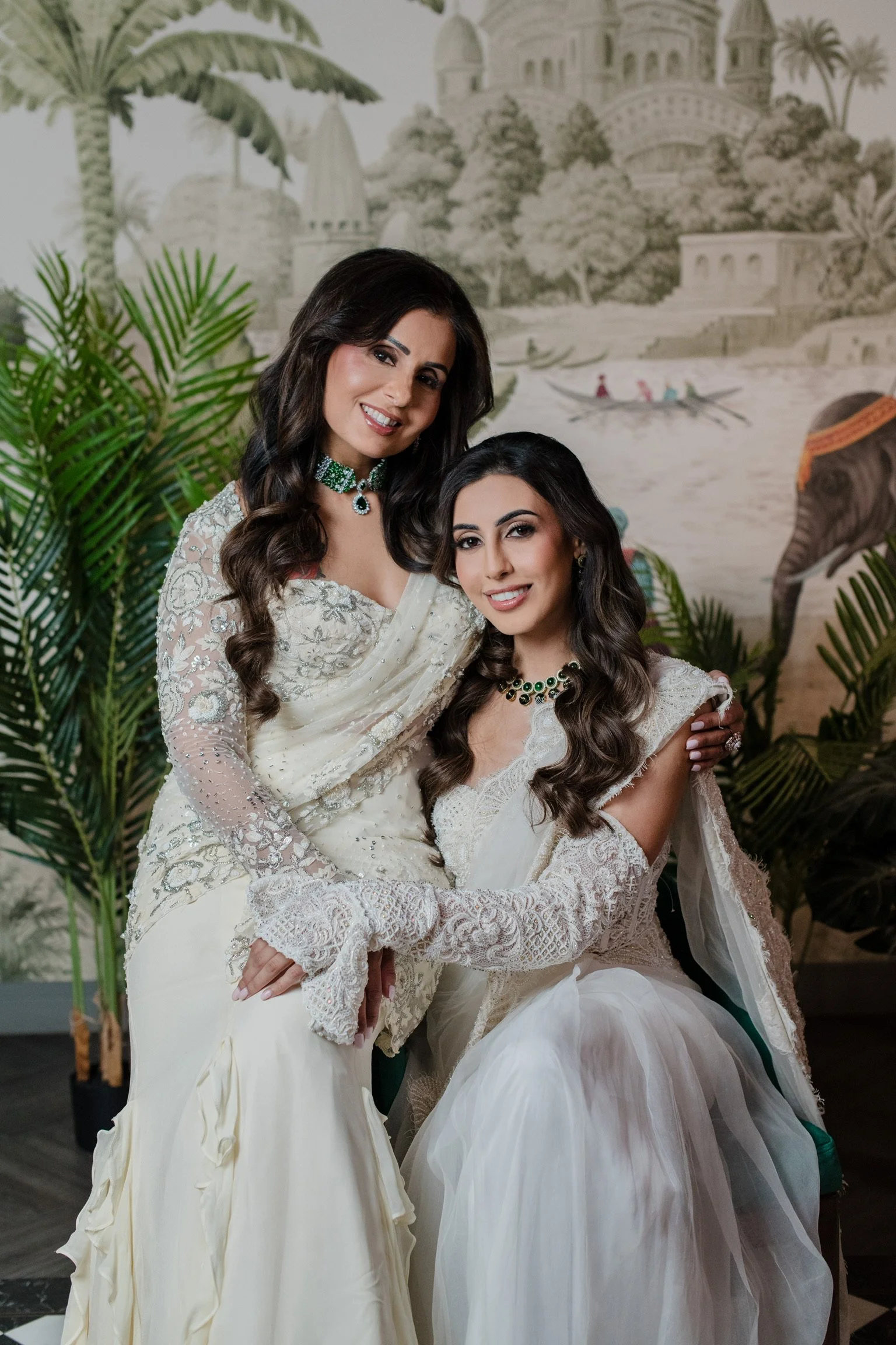 Two women dressed in elegant white gowns, smiling and embracing each other in front of a painted background depicting a castle, palm trees, and a boat on water.