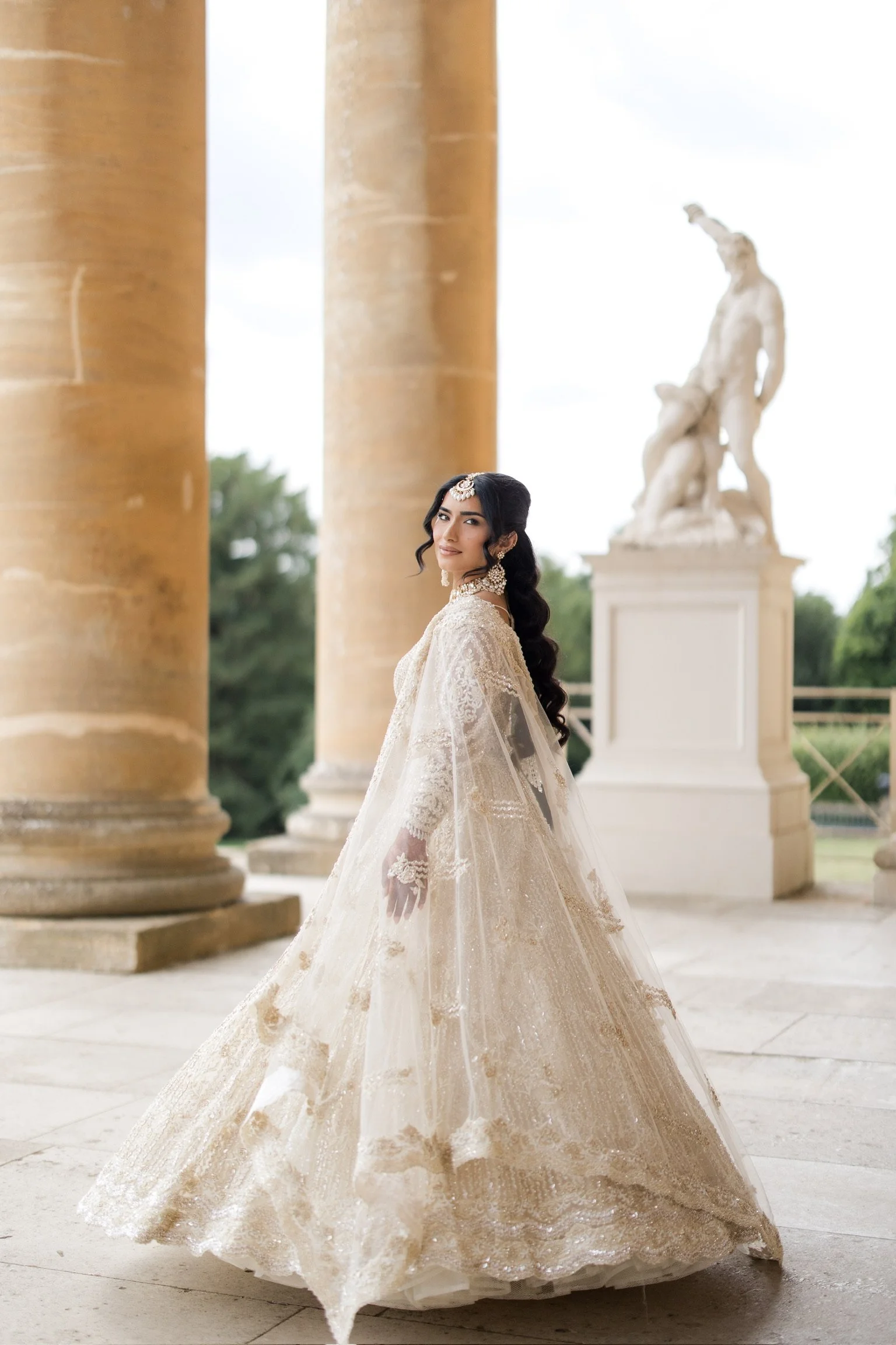 A woman in an ornate cream-colored gown with embroidery and lace details, standing outdoors near large columns with a classical sculpture in the background.