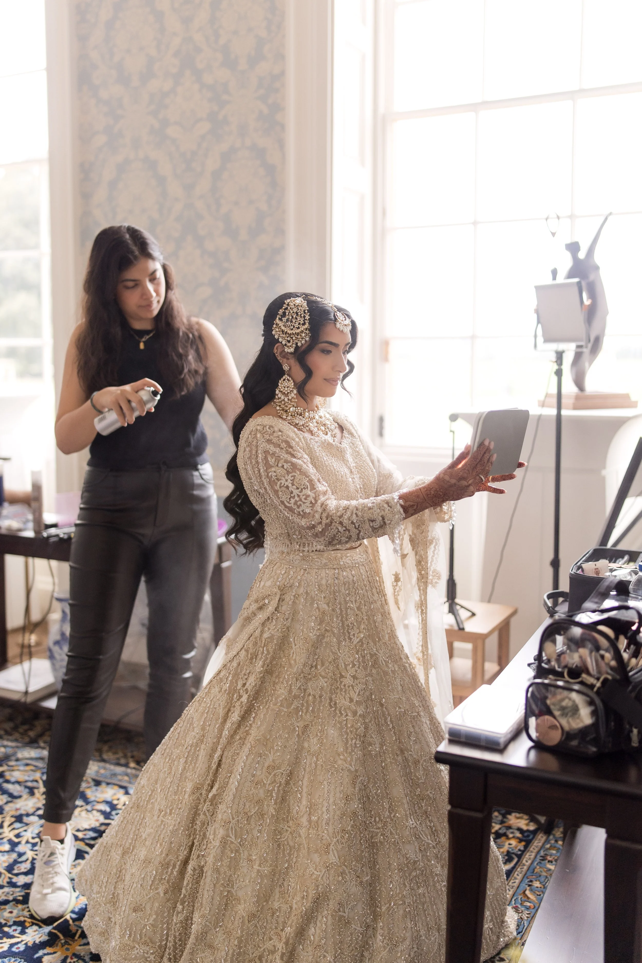 A bride dressed in a traditional, ornate beige lace wedding gown with jewelry and hair accessories, is looking at a tablet while a hairstylist stands behind her, spraying her hair. The room is well-lit with large windows, and there is a decorative sc