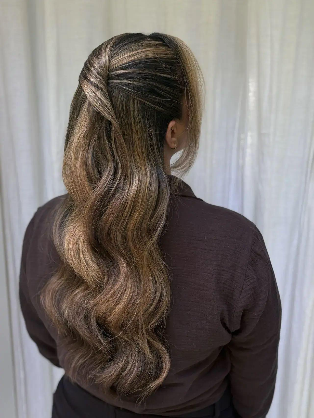 The image shows a woman with long, wavy hair that is styled with a twist at the crown, with the rest of her hair cascading down her back. She is wearing a dark brown top, and her face is turned away from the camera, showing her profile.
