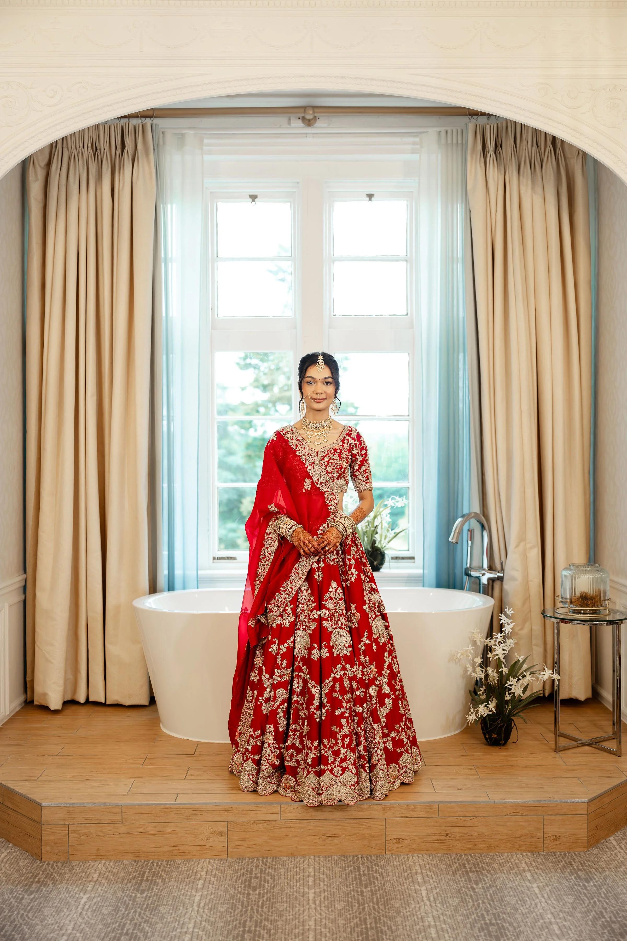 A woman dressed in a red and gold traditional dress standing by a bathtub in a well-lit room with large window, cream-colored curtains, and decorative accents.