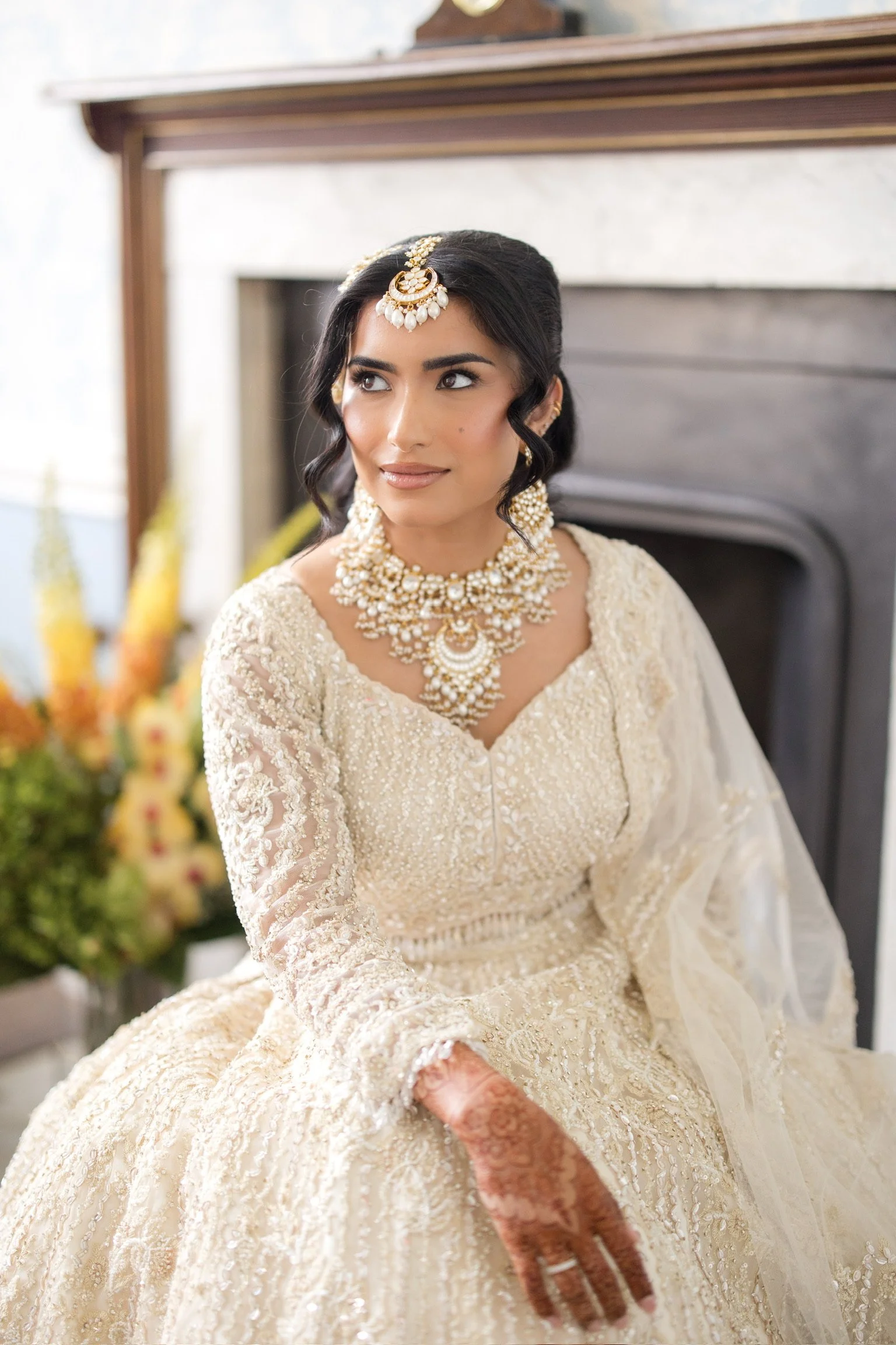 A woman dressed in an elegant cream-colored traditional dress, adorned with intricate lace and embroidery, wearing elaborate jewelry including a headpiece, necklace, earrings, and rings, sitting beside a fireplace with a blurred background of flowers