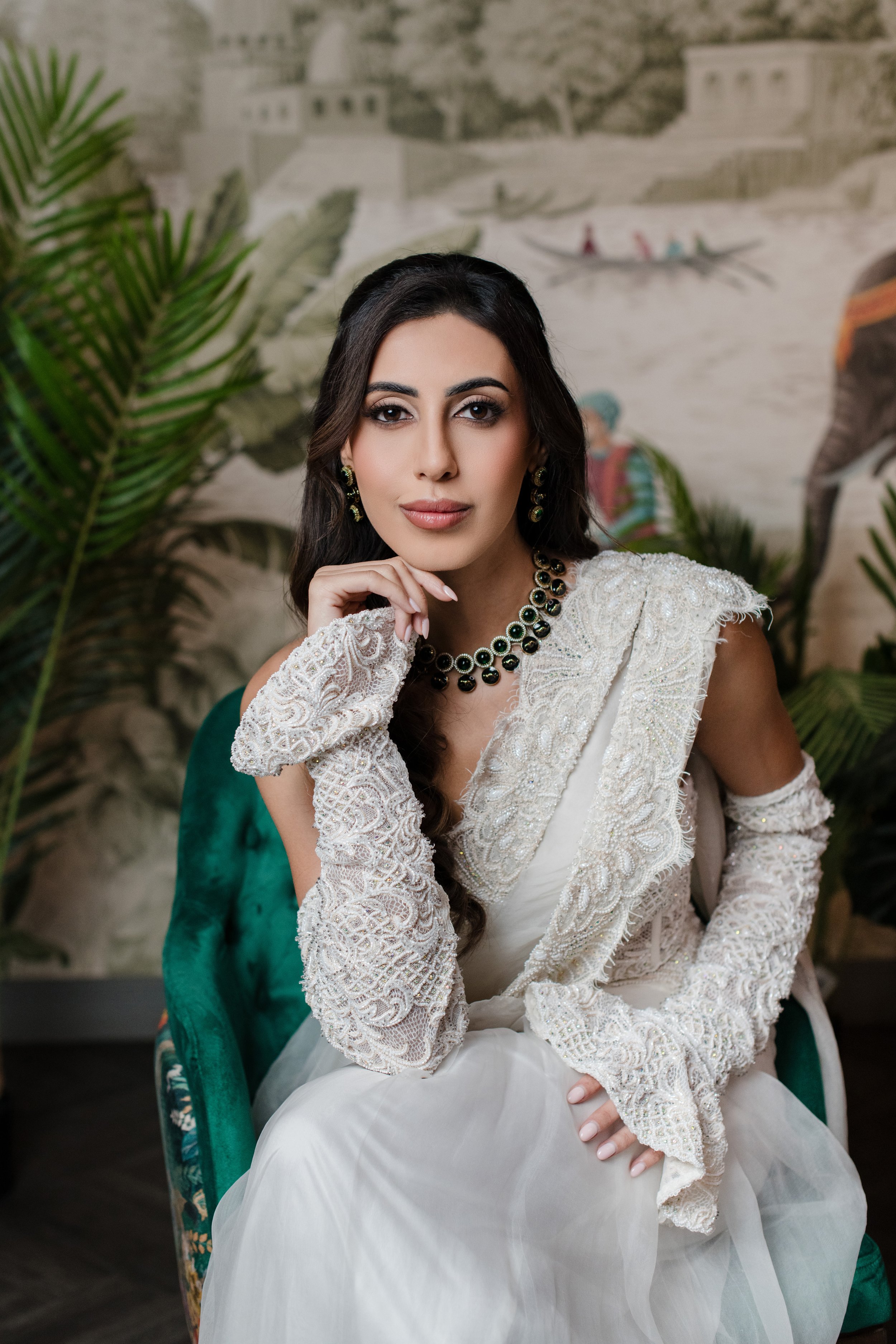 A woman in traditional Indian attire, wearing a white lace dupatta and a cream-colored saree, sitting on a green velvet chair with her hand resting on her chin. She is dressed with jewelry, including a black and gold necklace and matching earrings. In the background, there is a mural of a historical site with elephants and a boat, along with green plants.