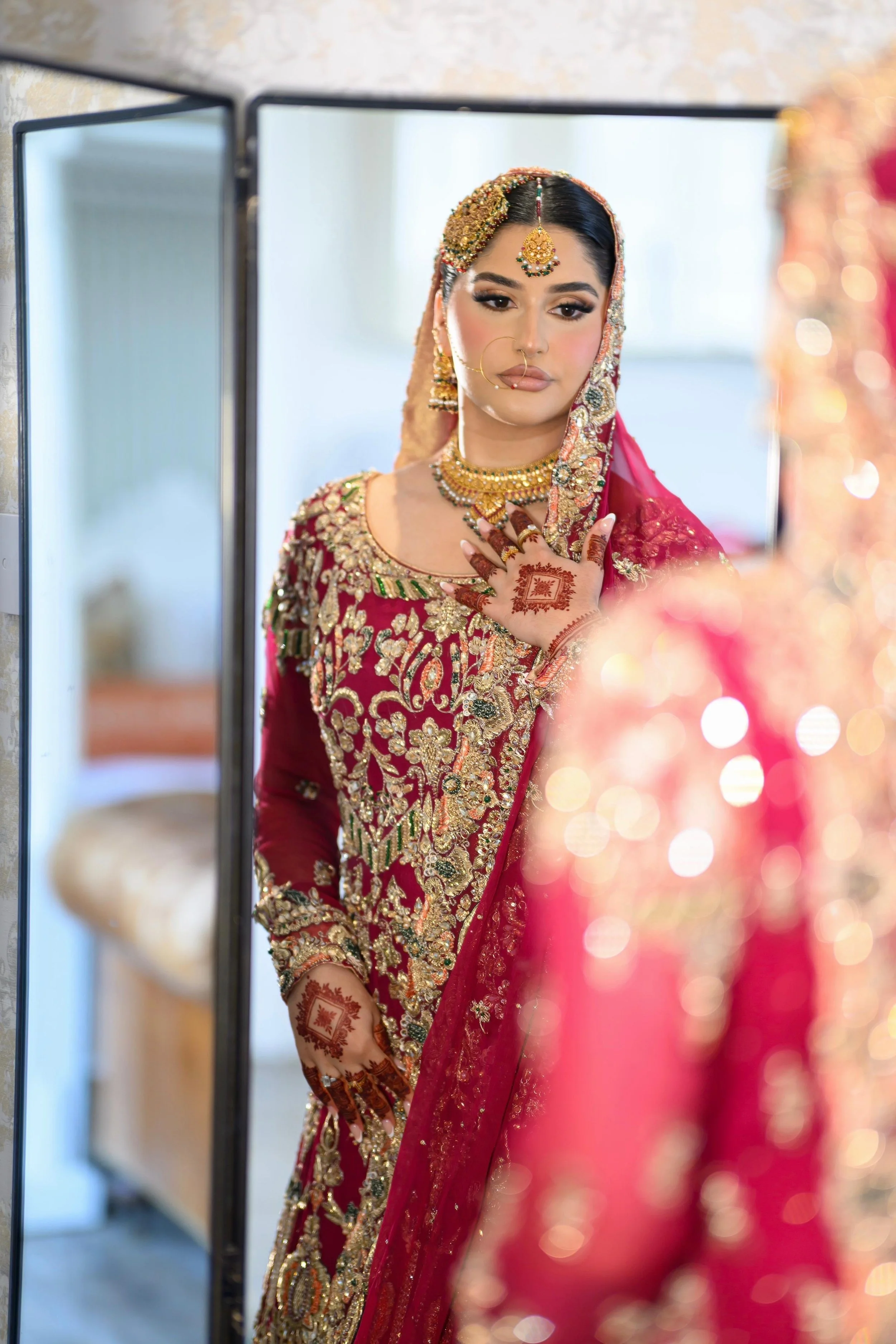 A woman in traditional Indian bridal attire with ornate gold jewelry, henna designs on her hands, and a red embroidered saree, looking at herself in a mirror.
