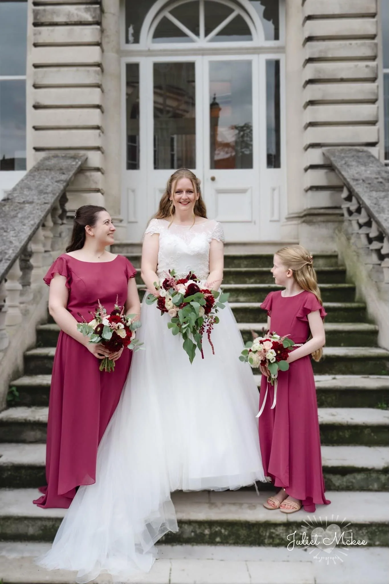 A bride and two bridesmaids, all smiling and holding bouquets, standing on outdoor stone staircase in front of a building with large white windows and a door.
