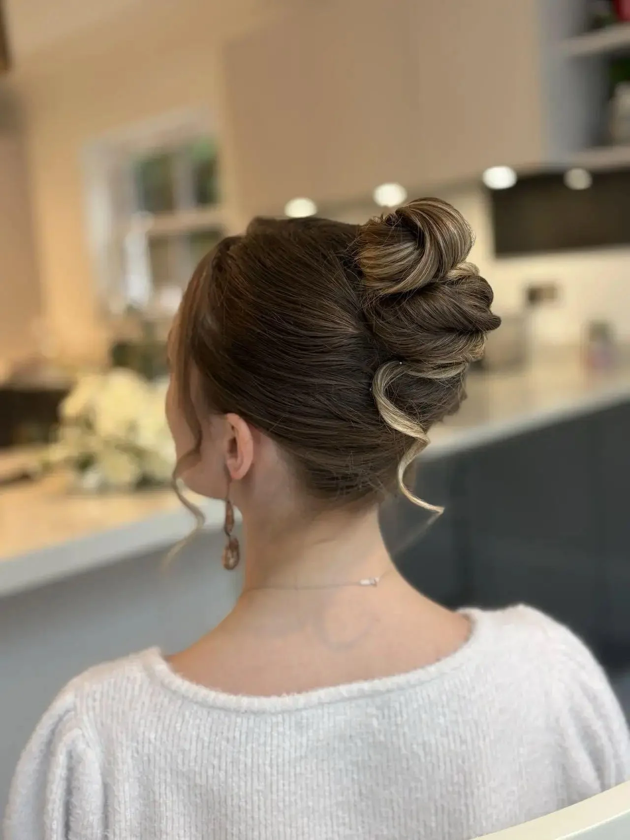 A woman with styled brown hair in an elegant updo with curls, wearing a white sweater and earrings, in a room with blurred background.