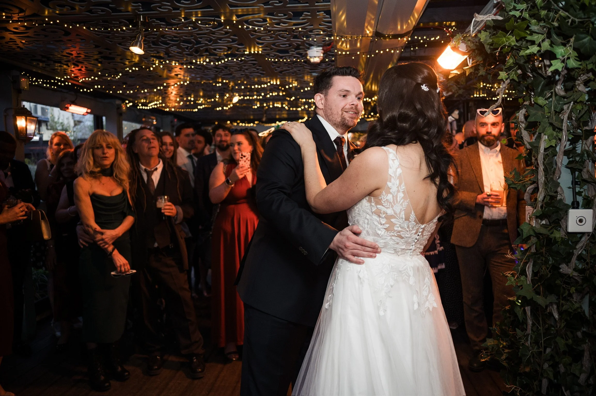 A bride and groom dancing at their wedding reception, surrounded by guests under string lights and a decorated ceiling.