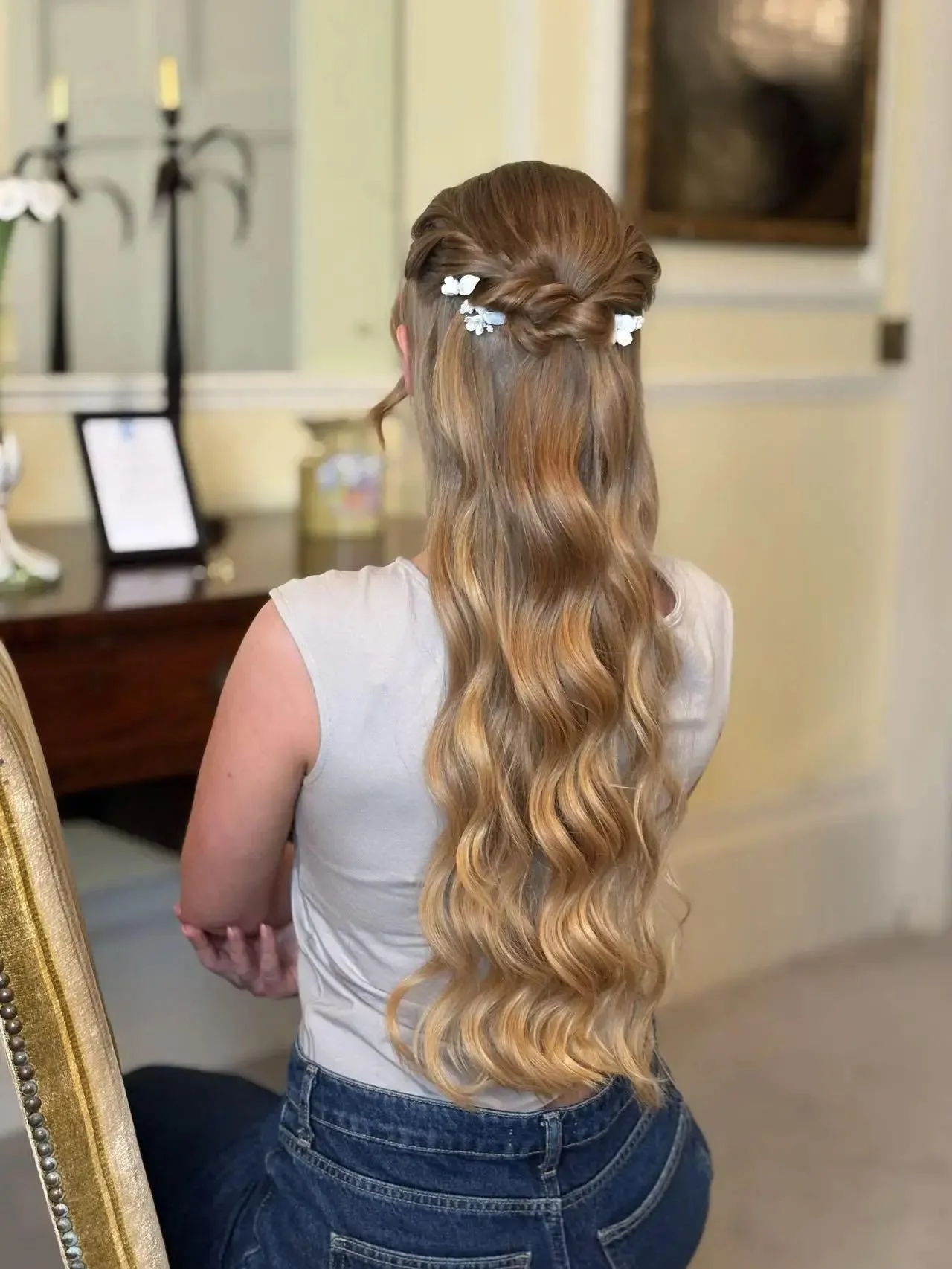 Woman with long, wavy, blonde hair styled with a twisted crown braid adorned with white flowers, sitting indoors with her back to the camera.
