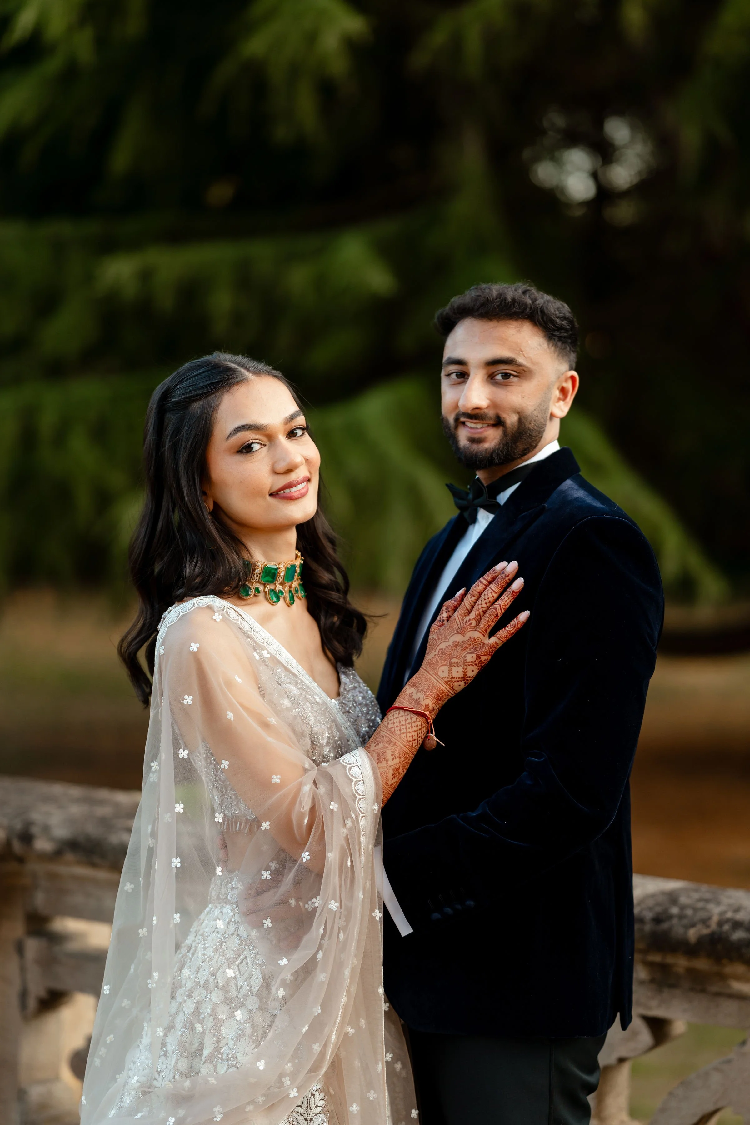 A newlywed couple standing outdoors, the woman in a white wedding dress and the man in a black tuxedo with a bowtie, surrounded by green trees.