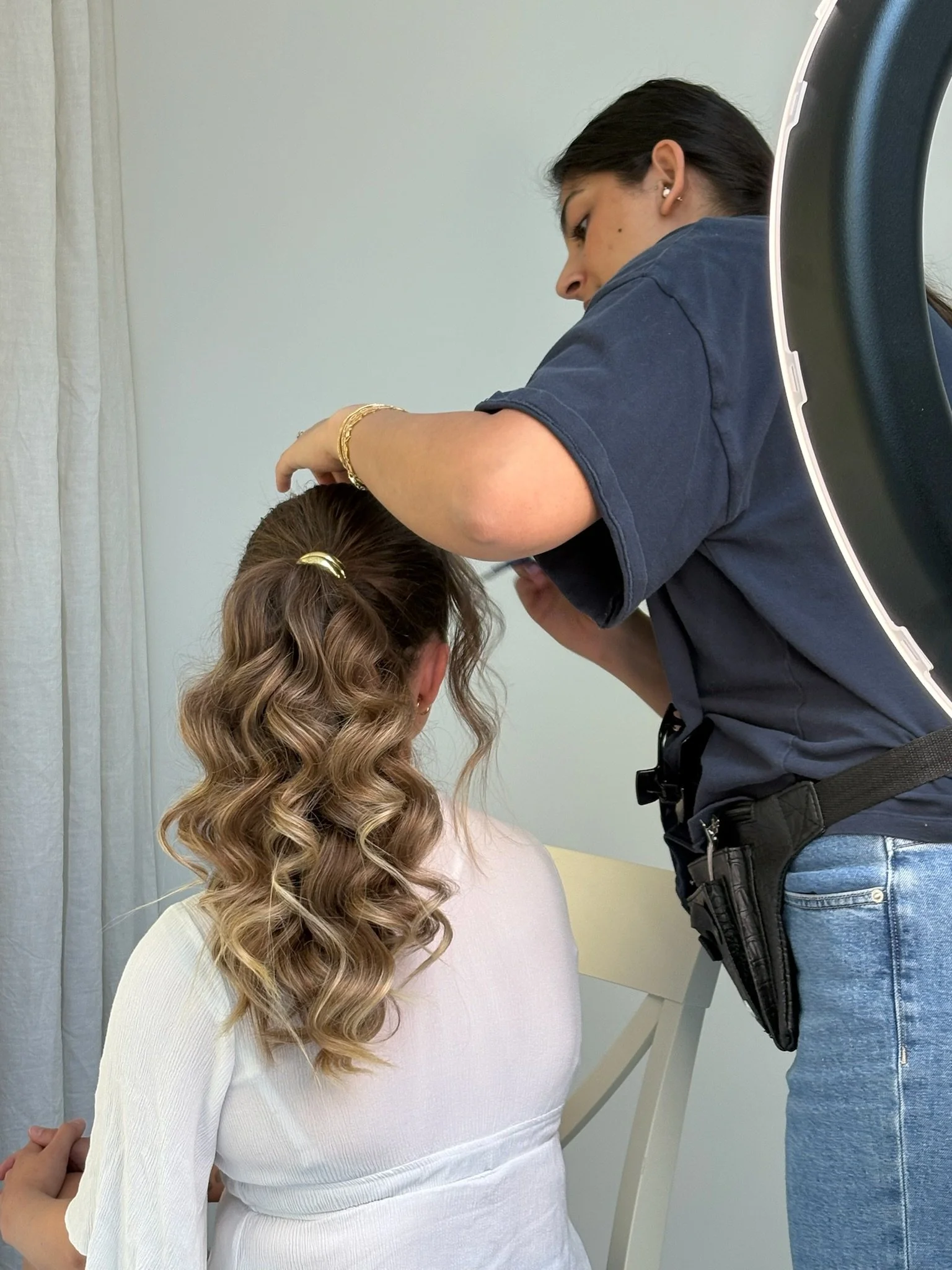 A person with dark hair, wearing a dark t-shirt and blue jeans, working on hair styled in loose curls, while another person, with light brown hair in a ponytail and wearing a white top, stands in front of them. The scene appears to be in a salon or similar setting.