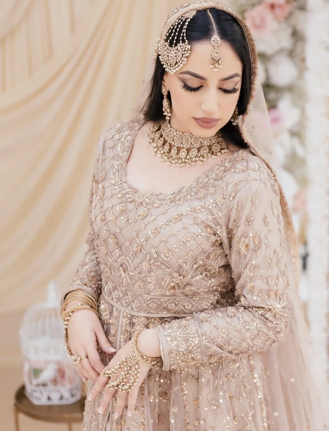 A woman dressed in an ornate, blush pink traditional South Asian bridal outfit, wearing extensive gold and pearl jewelry, standing against a beige and floral background.