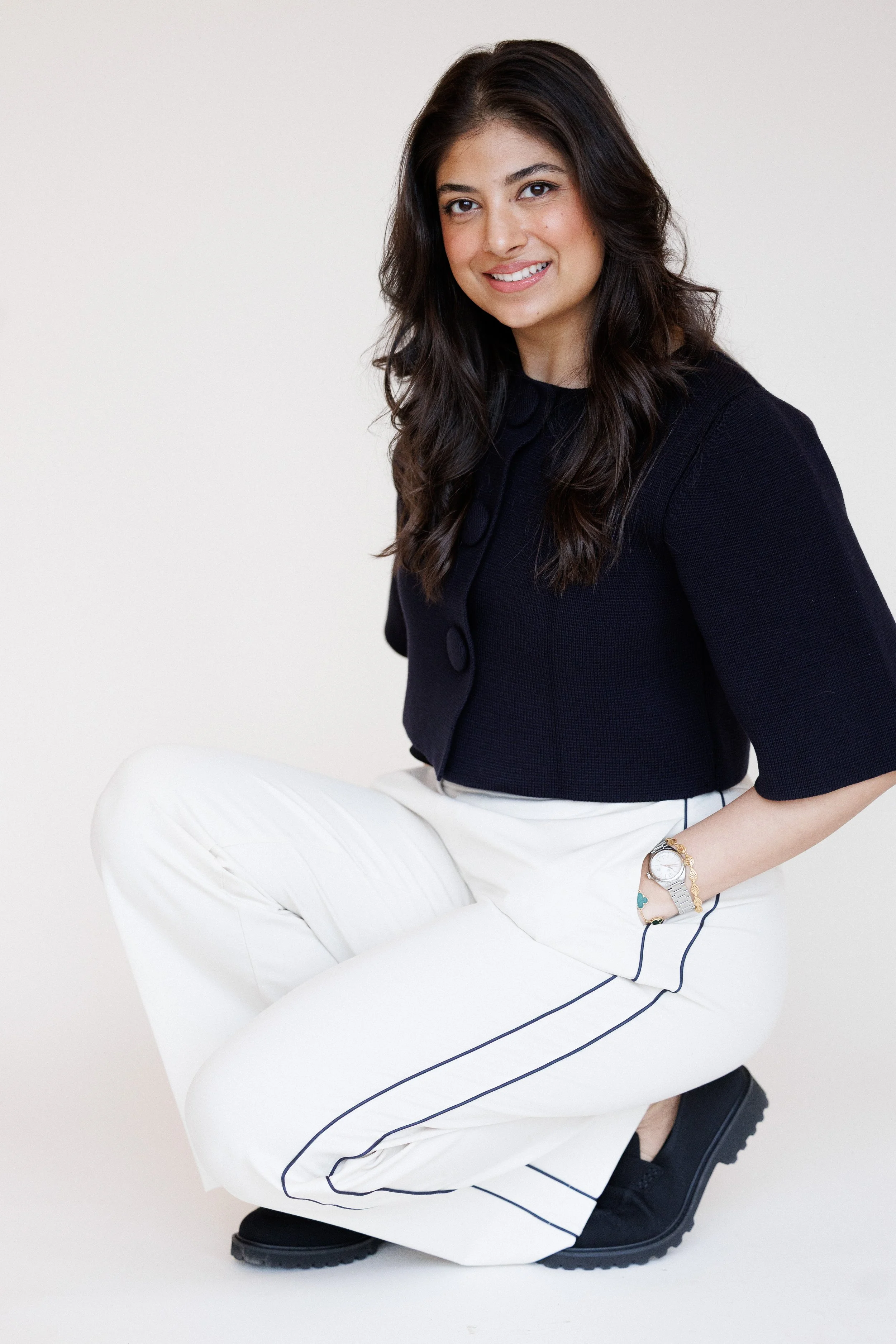 A woman with long dark hair, wearing a black top and white pants with black piping, sitting with one knee up on a white chair, smiling at the camera.