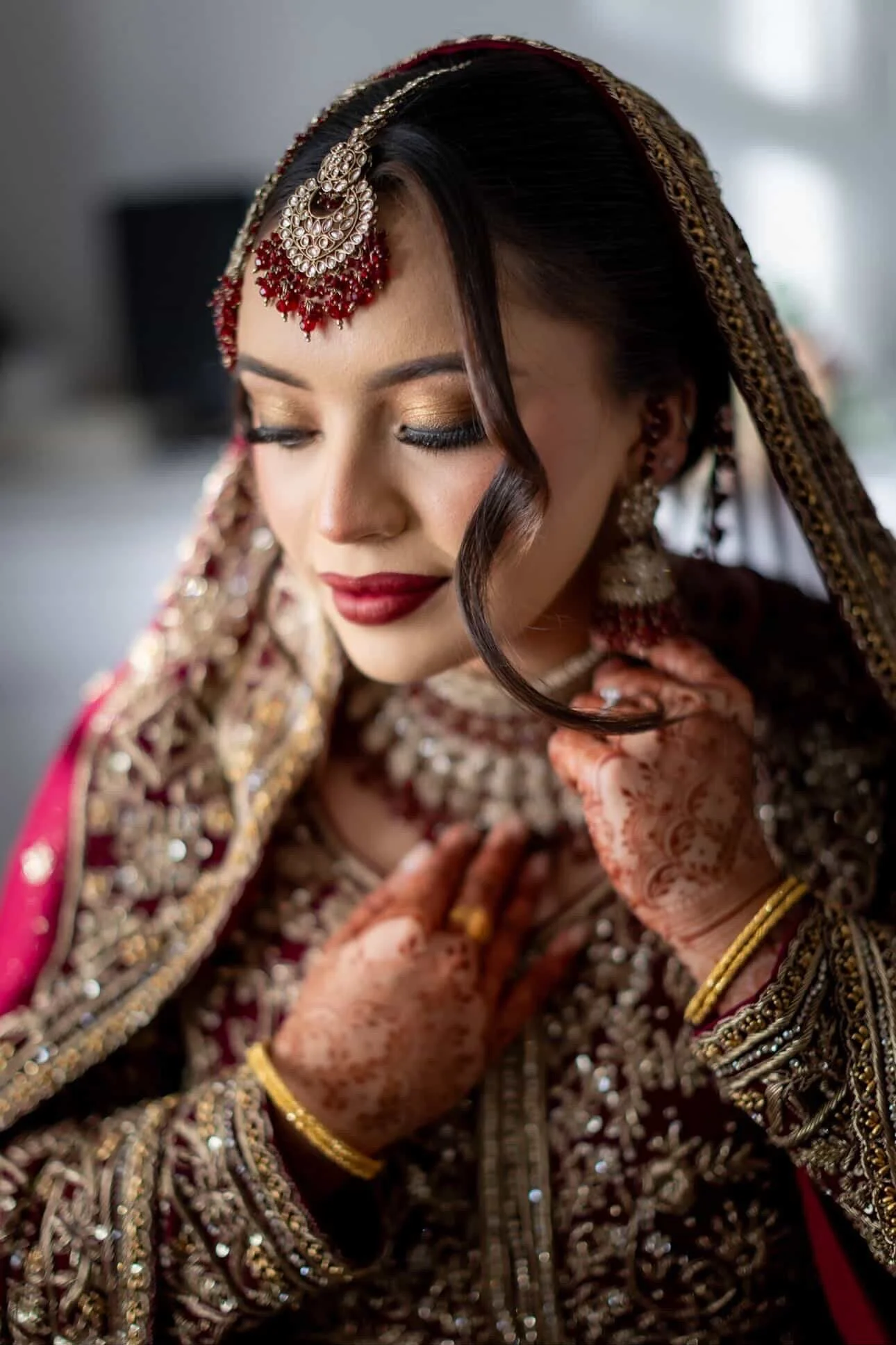 Close-up of a woman dressed in traditional Indian wedding attire, with intricate jewelry and henna on her hands, looking down with a gentle smile.