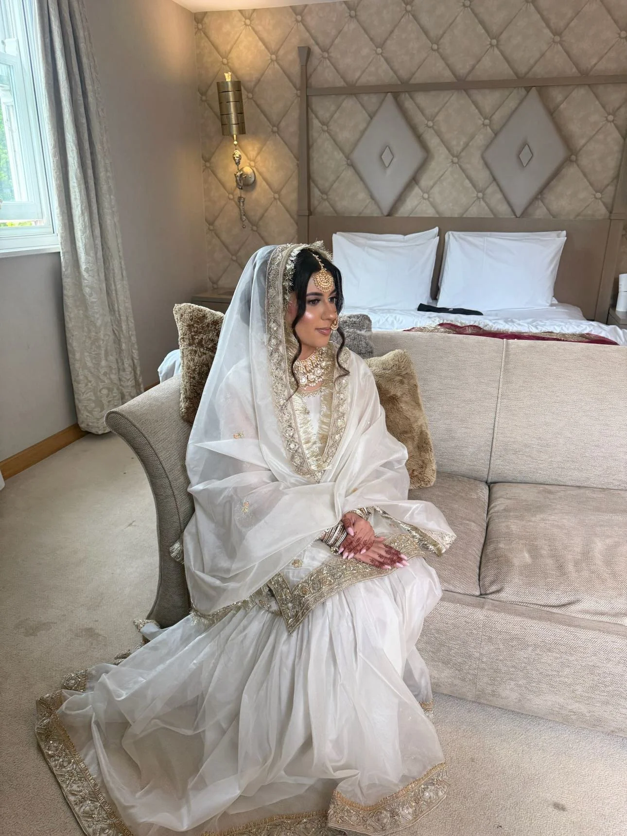 A woman dressed in traditional wedding attire, sitting on a beige sofa in a decorated room. She wears a white embroidered dress with gold accents, a matching headscarf, and jewelry including a necklace and forehead ornament. Her hands are folded on h