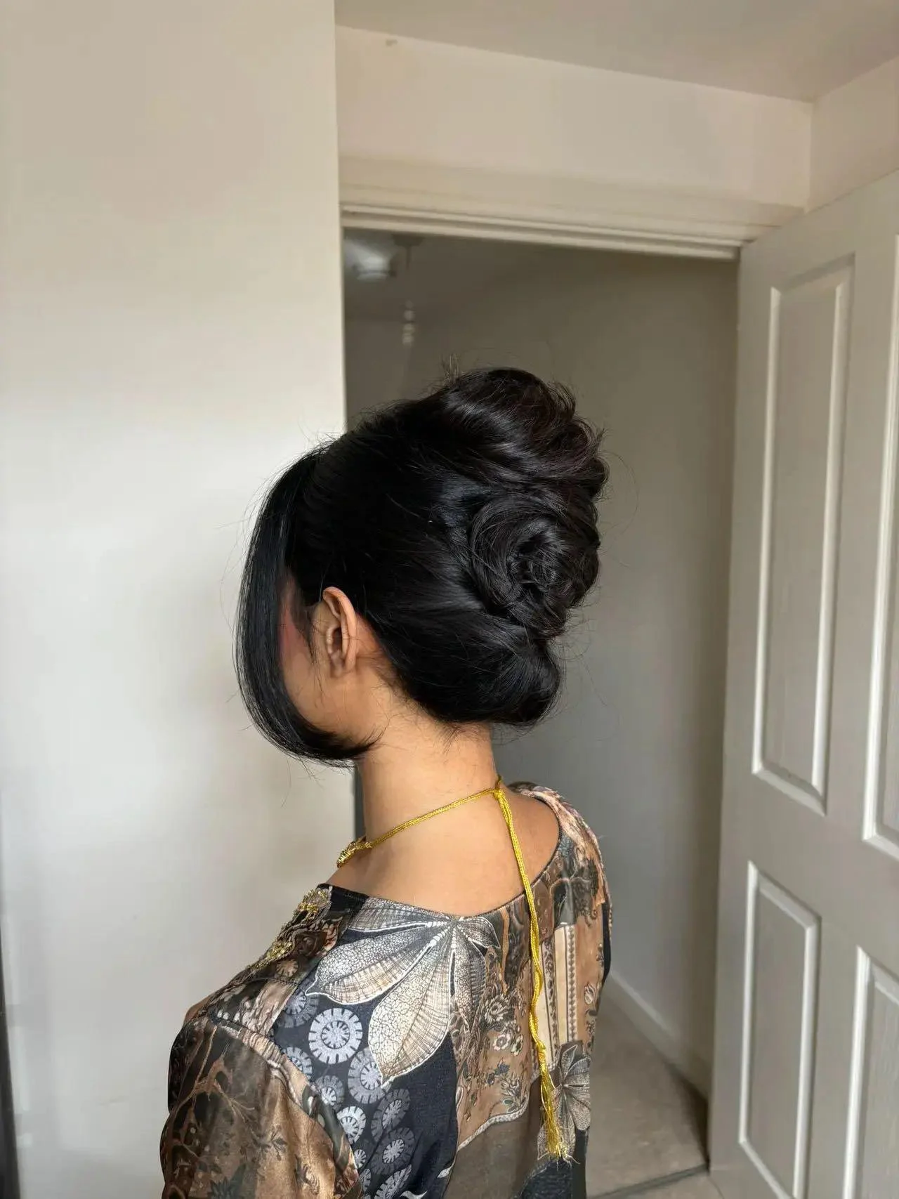 A woman with dark hair styled in an elegant updo, wearing a patterned dress and a gold necklace, standing in a room with a closed white door and beige walls.