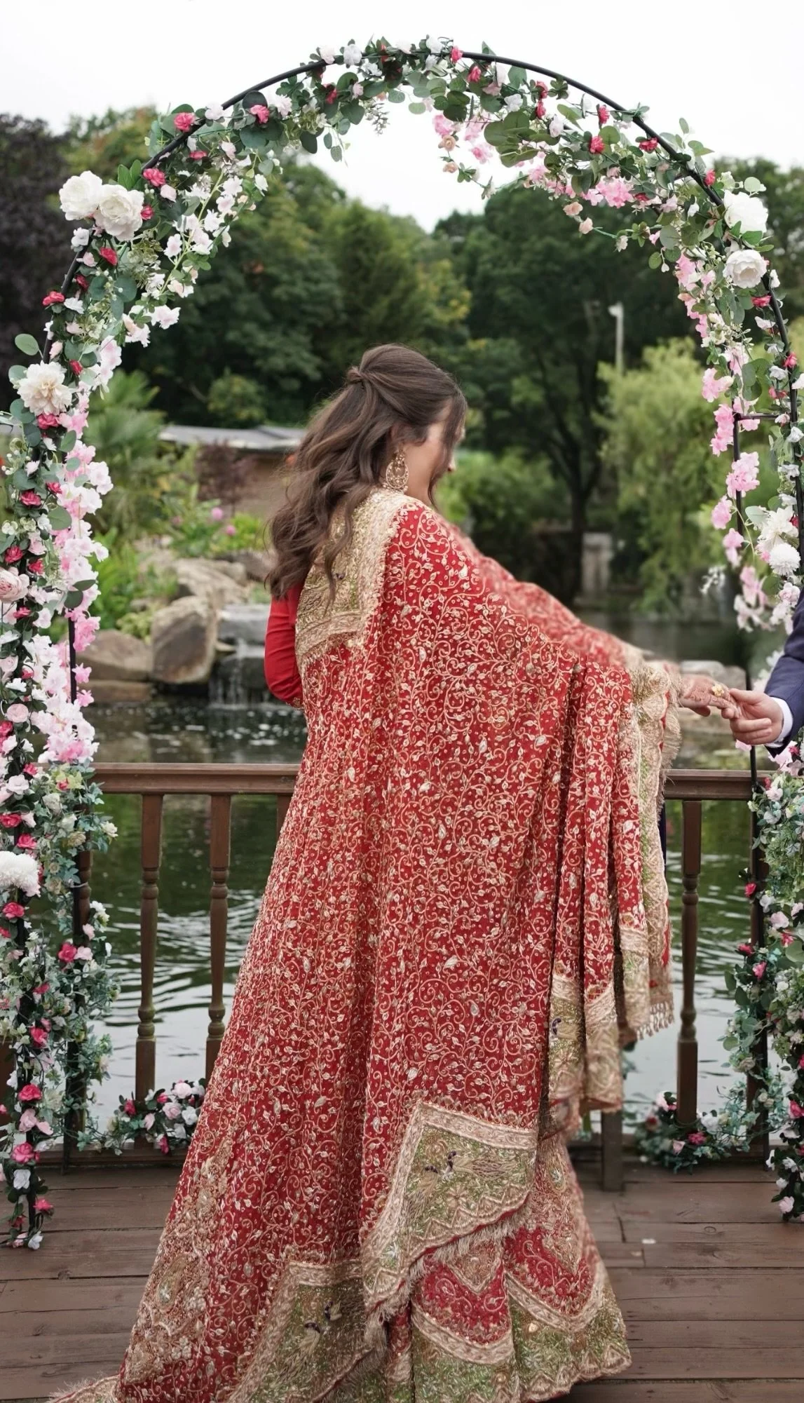 A woman dressed in an ornate red and gold traditional dress at a wedding or celebration, standing under a floral arch by a lake.