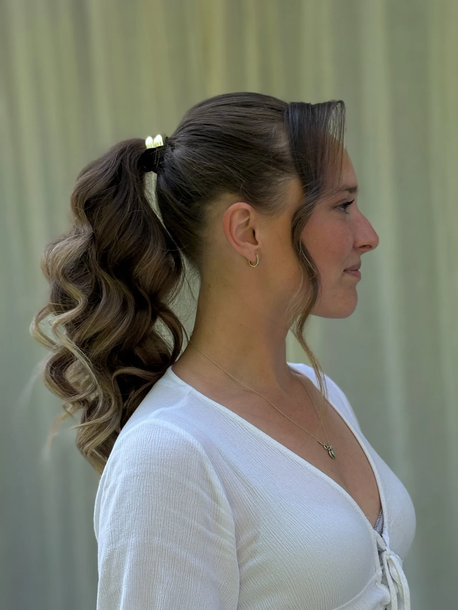 Profile view of a woman with long, brown, wavy hair tied back with a white hair tie, wearing a white top with a V-neck and lace-up front, and gold earrings and necklace, standing outdoors with a blurred green background.