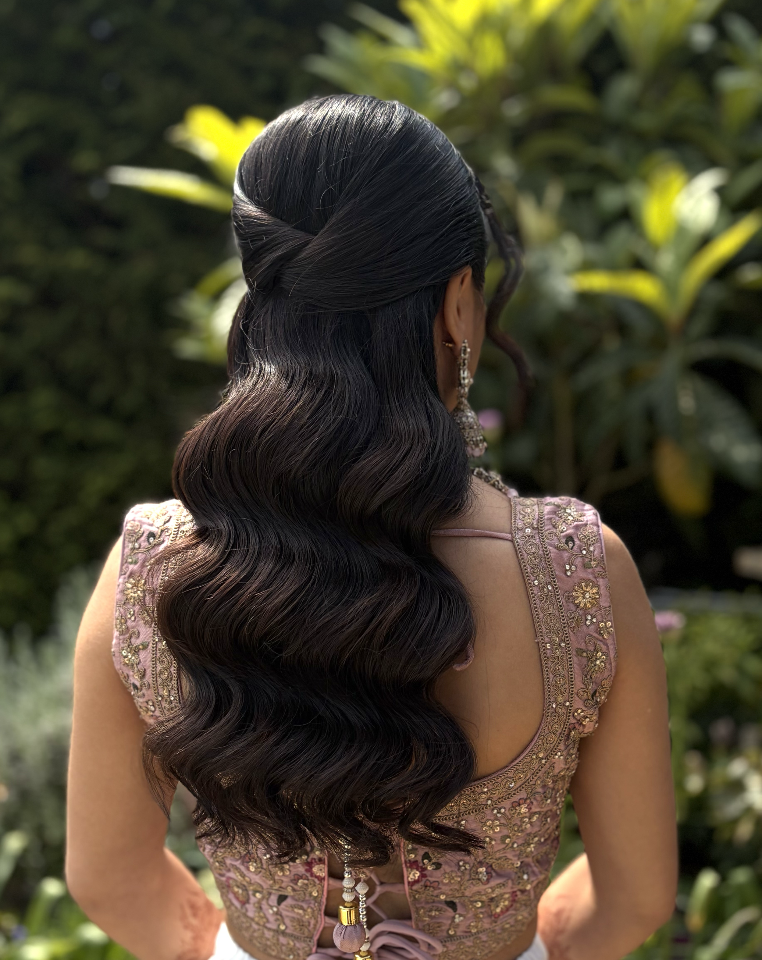 A woman with long, dark, wavy hair styled in loose waves, wearing earrings and a pink, embroidered traditional Indian blouse with detailed gold and silver embellishments, standing outdoors against a blurred background of green foliage.