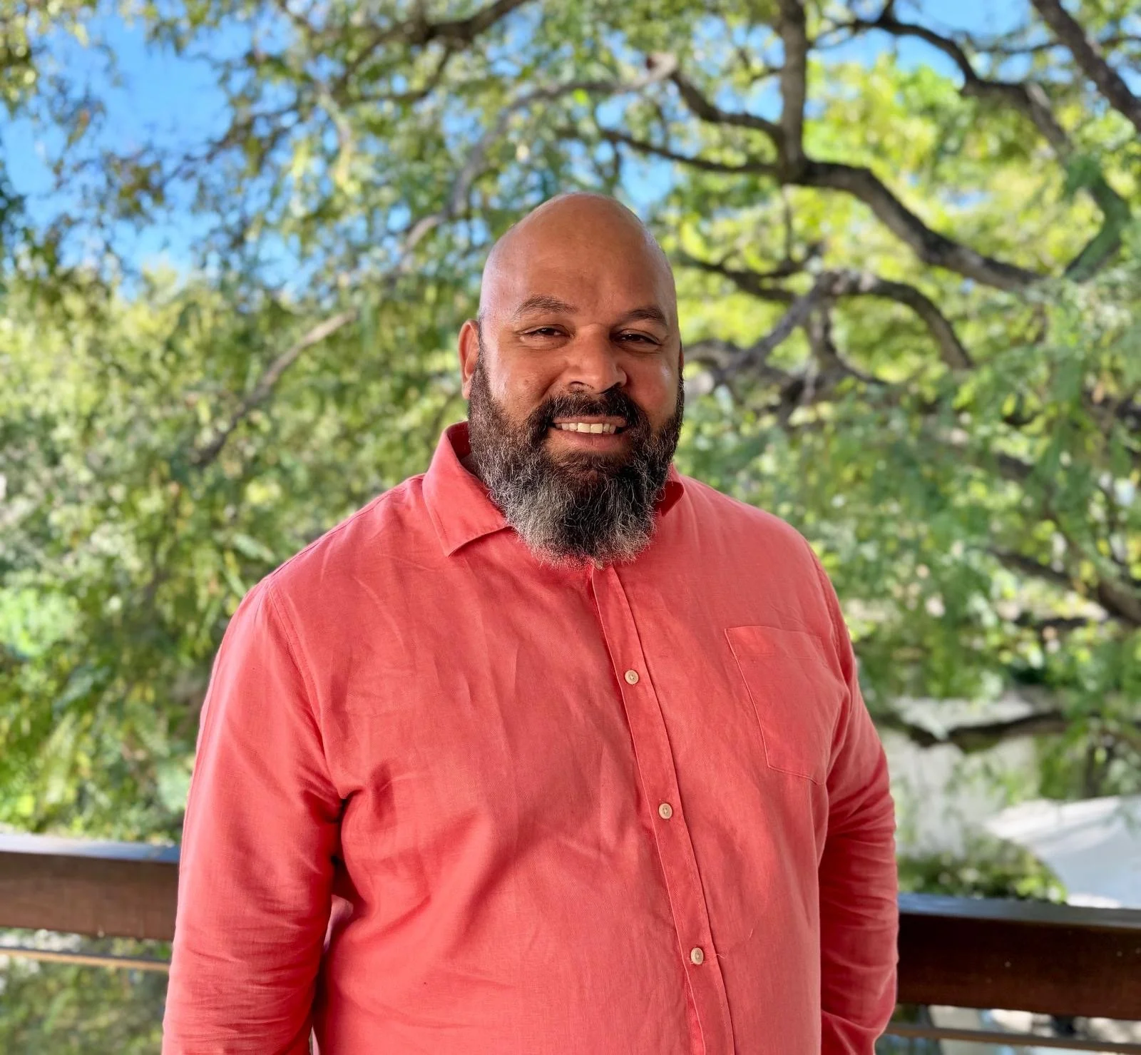 A man with a beard wearing a salmon-colored button-up shirt, standing outdoors with green trees and blue sky in the background.