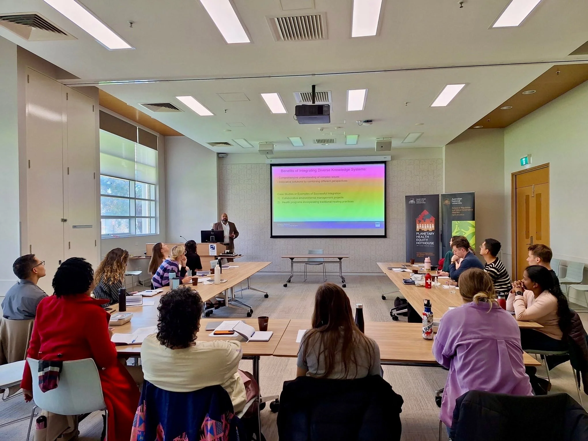 A classroom or conference room with numerous students seated around U-shaped tables listening to a speaker at the front. The speaker stands near a large screen displaying a presentation. The room has large windows, ceiling lights, and banners on the side walls.