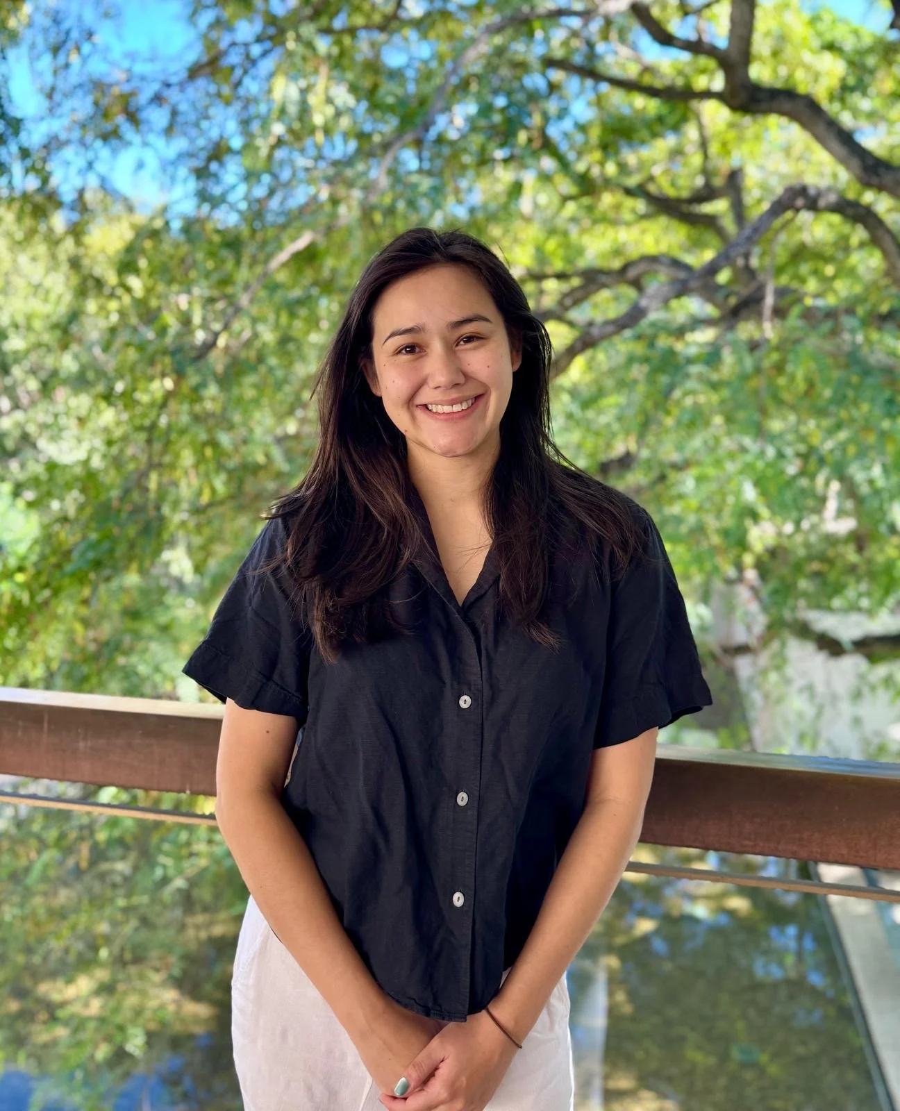 A young woman with long dark hair, smiling and wearing a black button-up shirt, standing on a wooden balcony surrounded by green trees and foliage.