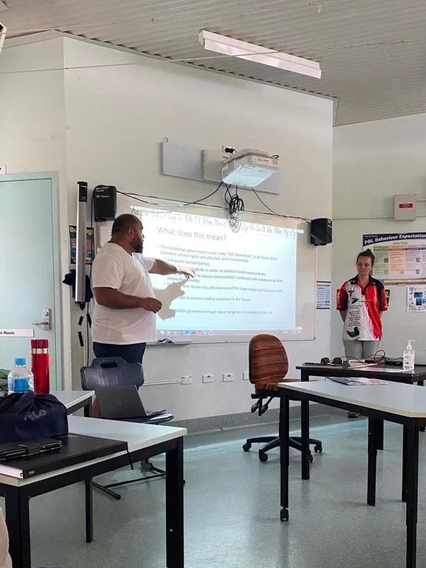 A man is standing and pointing at a whiteboard in a classroom or training room, while a woman stands nearby with a red and black backpack, and there are tables, chairs, a hand sanitizer, and a water bottle in the room.