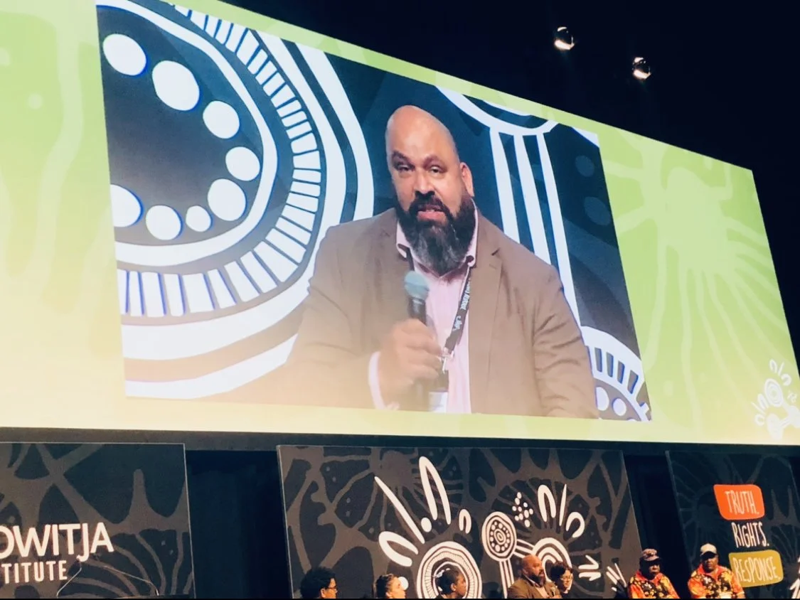 A man with a beard and bald head sitting on a panel, speaking into a handheld microphone. Behind him is a large screen showing his live feed, with colorful, abstract background patterns. Several people are seated at a panel table in front of the stage.
