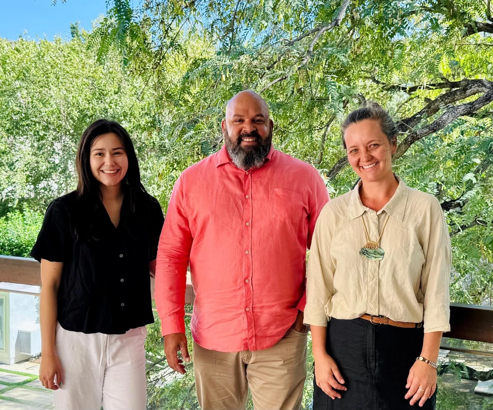 Three smiling adults standing outdoors on a deck with green trees in the background.