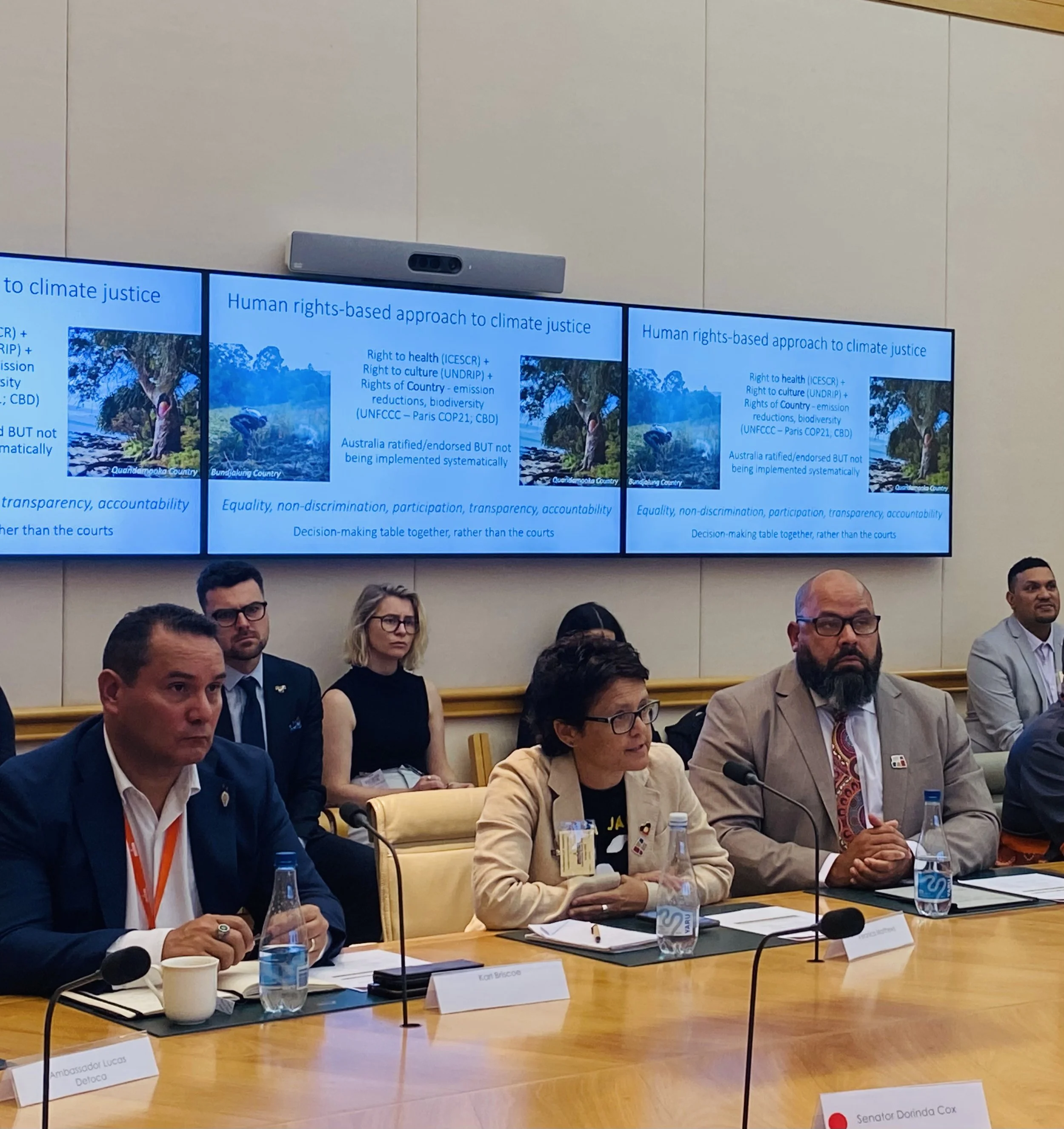 A diverse group of people seated at a conference table during a meeting or panel discussion. There are nameplates, water bottles, and microphones in front of each person. Behind the group, a screen displays a presentation about climate justice and human rights-based approaches to climate policy.