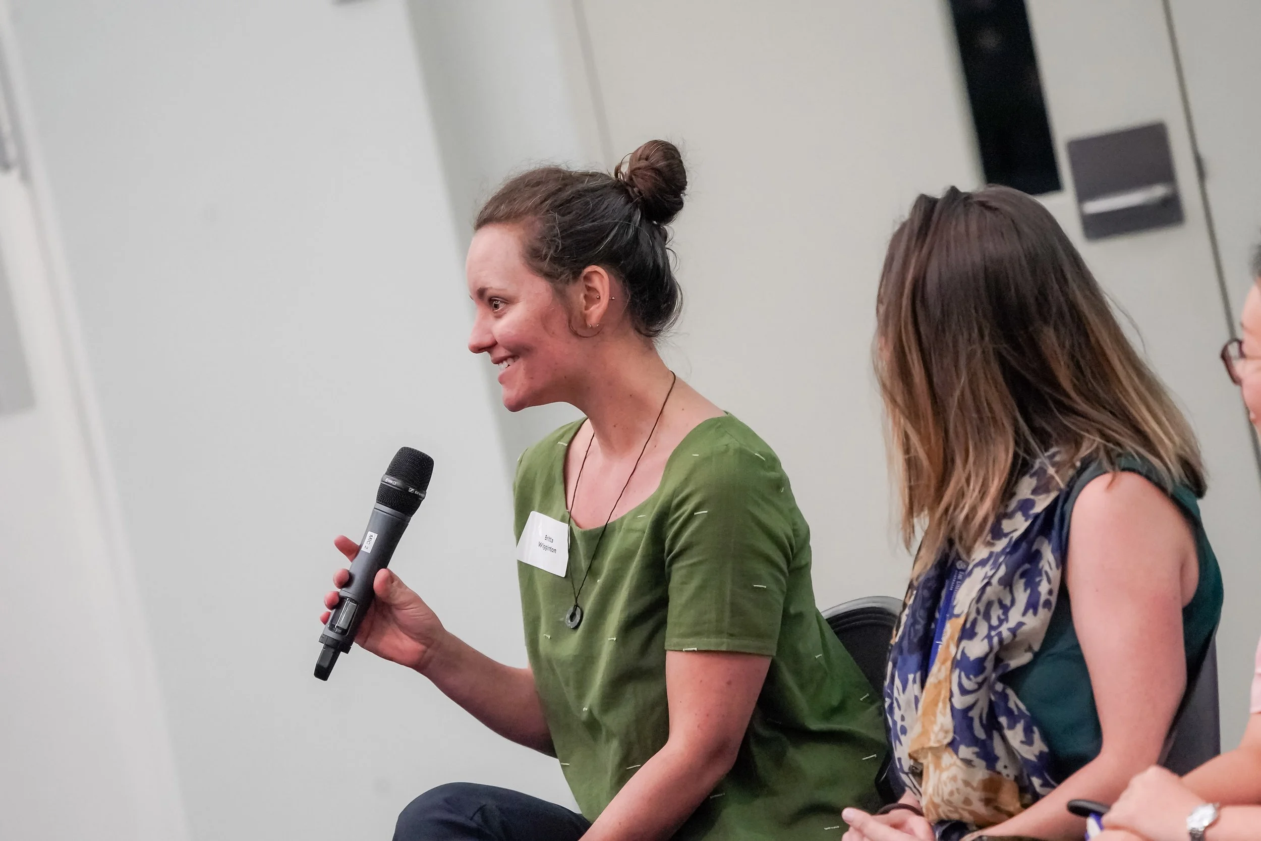 A woman speaking into a microphone during a panel discussion or presentation. She has brown hair tied up in a bun, and is wearing a green shirt with a name tag. Other people are seated next to her, listening.