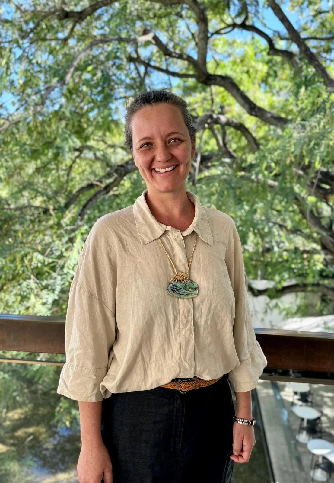A woman smiling outdoors with green trees in the background, wearing a beige blouse and a large pendant necklace.