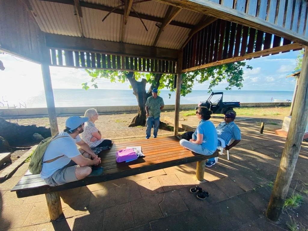 Group of six people sitting and standing under a wooden shelter by the beach, with one person speaking to the group. There are trees, a concrete barrier, and the ocean in the background.