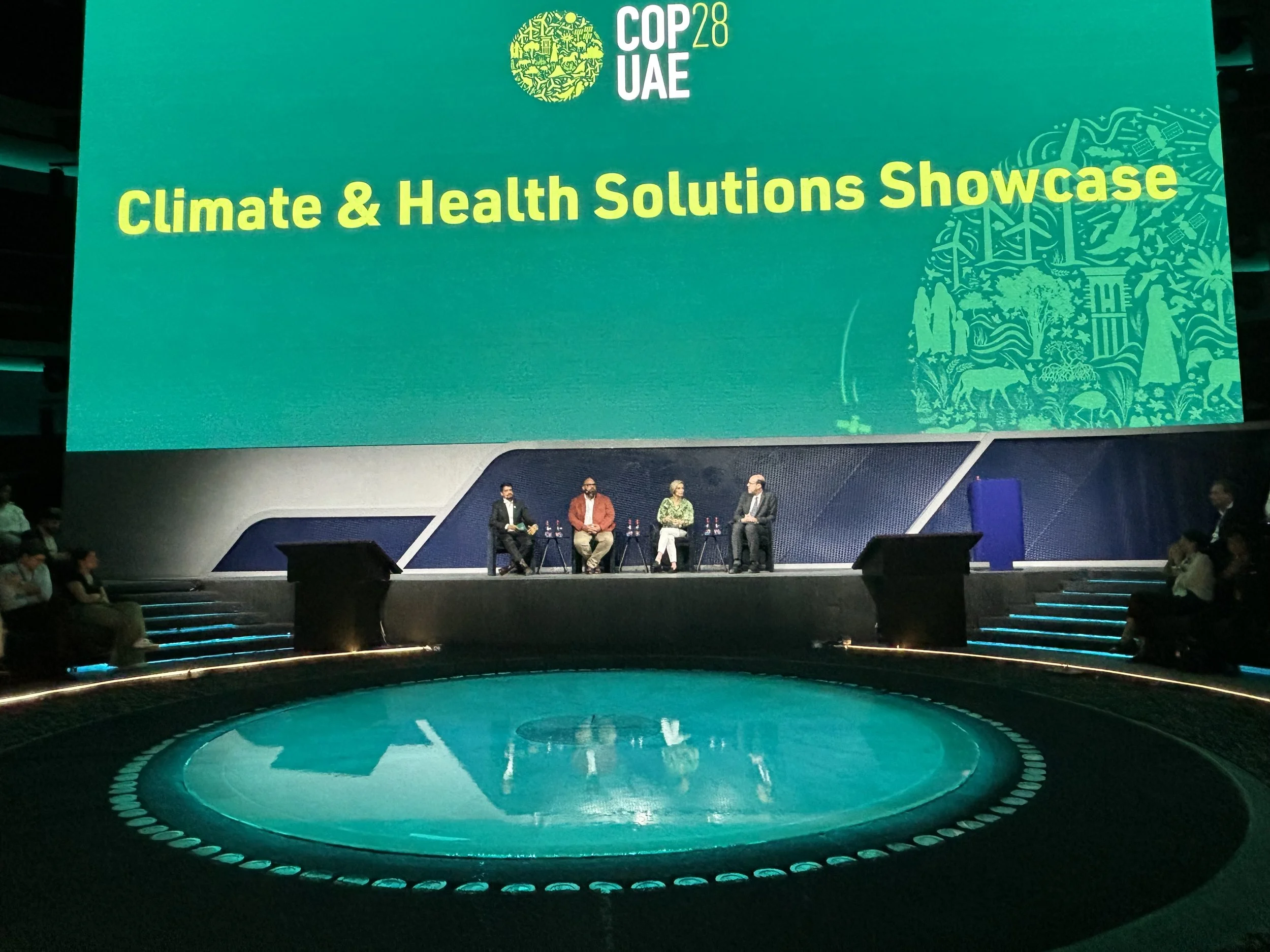 Large conference room stage with a big green screen displaying the words 'Climate & Health Solutions Showcase' and the logos 'COP28 UAE'. There are five people seated on stage and an audience in front.