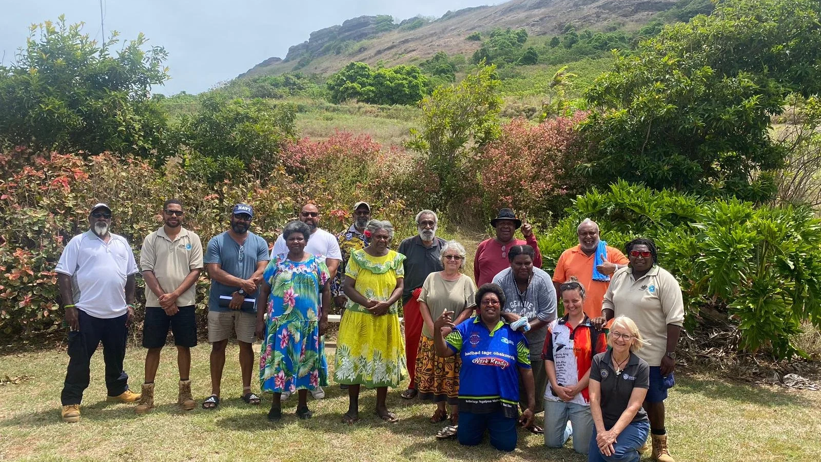Group of people standing outdoors on grass in front of lush greenery and mountains, posing for a photo on a sunny day.