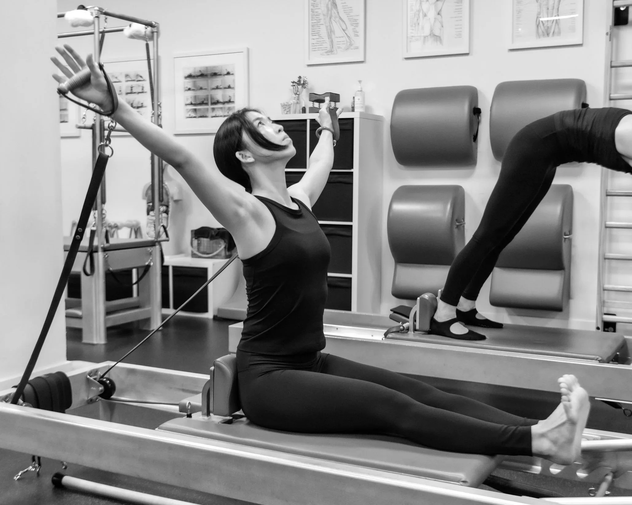 A woman practicing Pilates on a reformer machine in a gym, with her arms extended upward and head tilted back, and a mirror reflecting her pose.