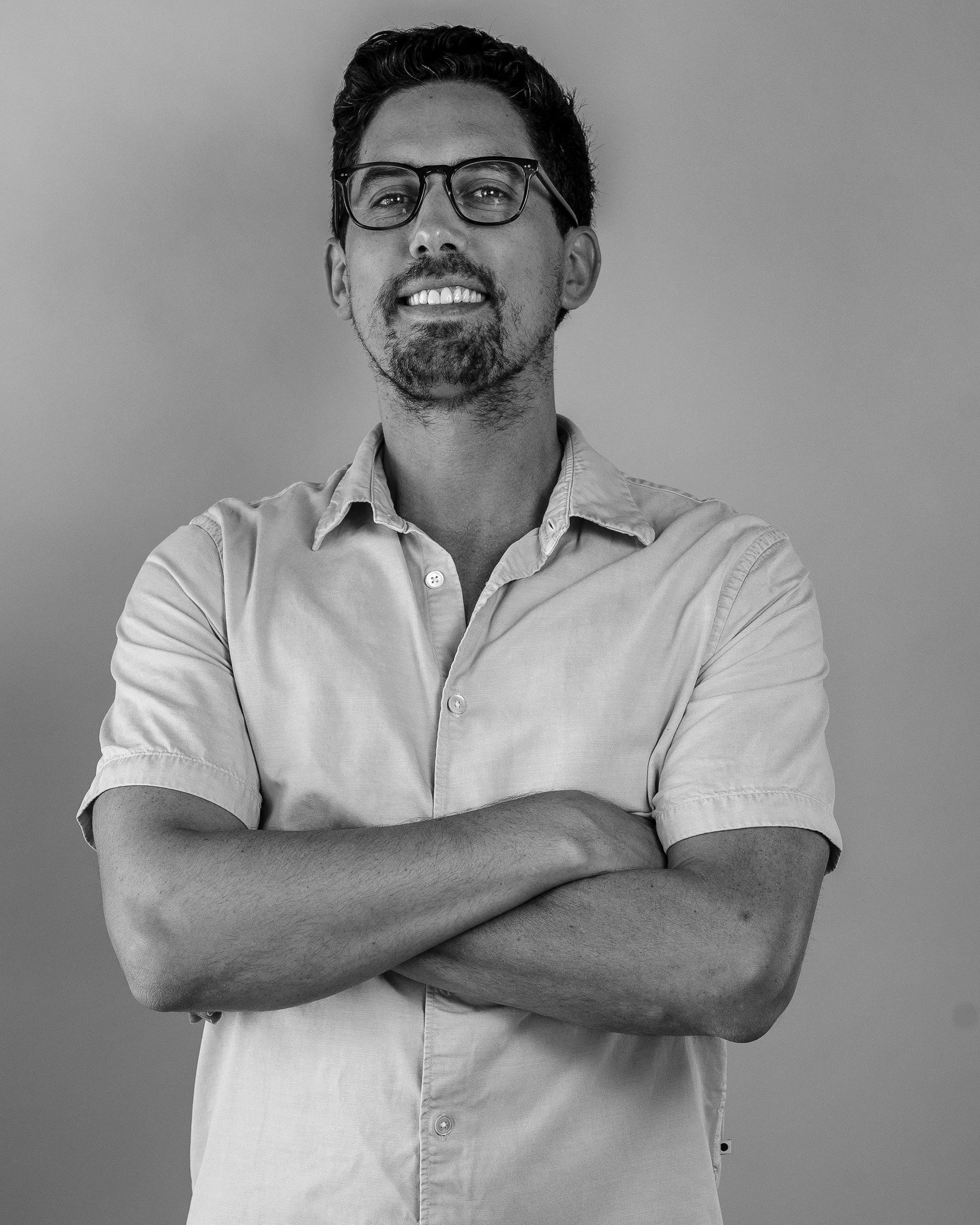 Black and white photograph of a man with glasses and facial hair, crossing his arms and smiling, wearing a button-up shirt, against a plain gray background.